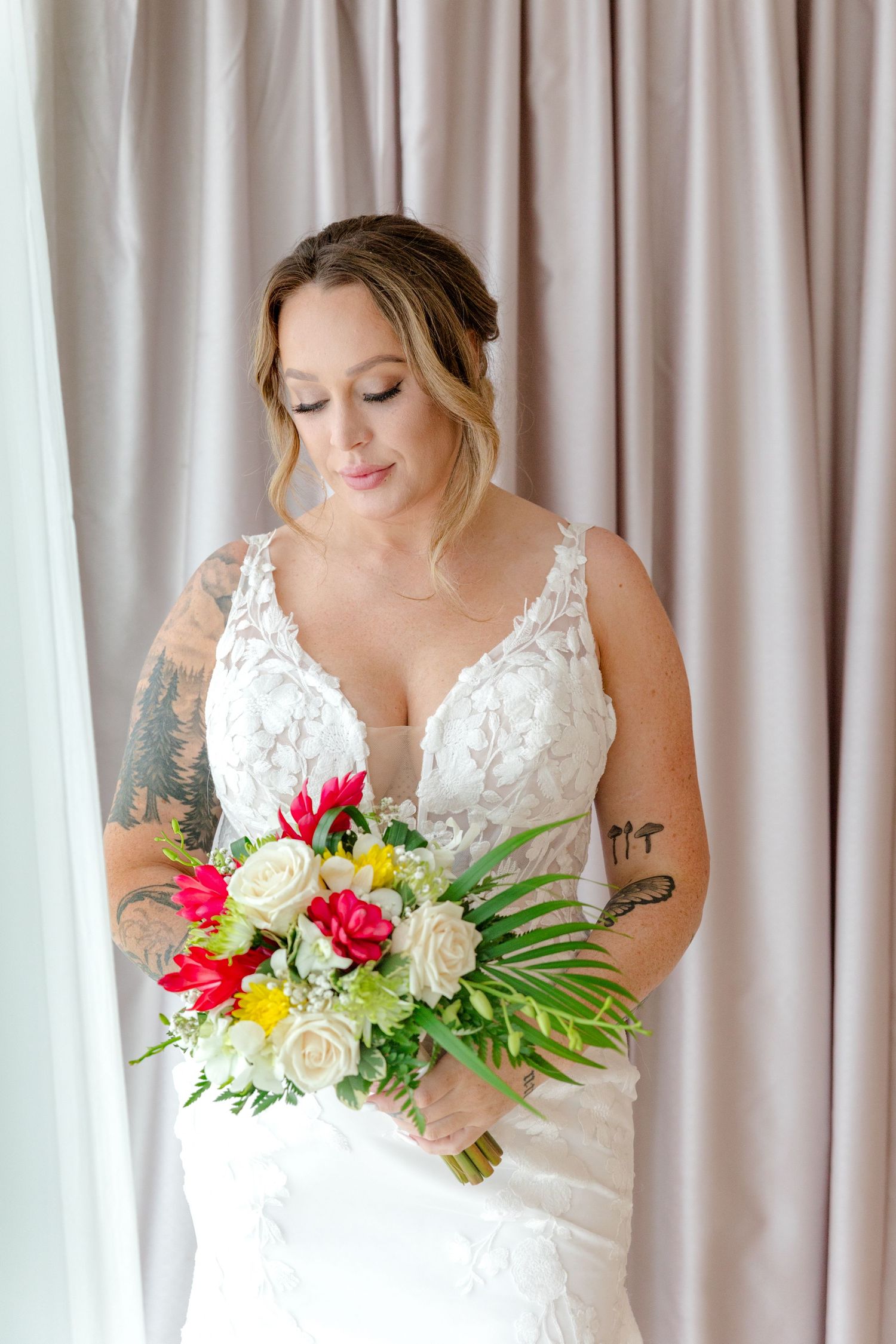 Tattooed bride in white lace gown holds colorful bouquet with red and cream flowers, looking down elegantly.