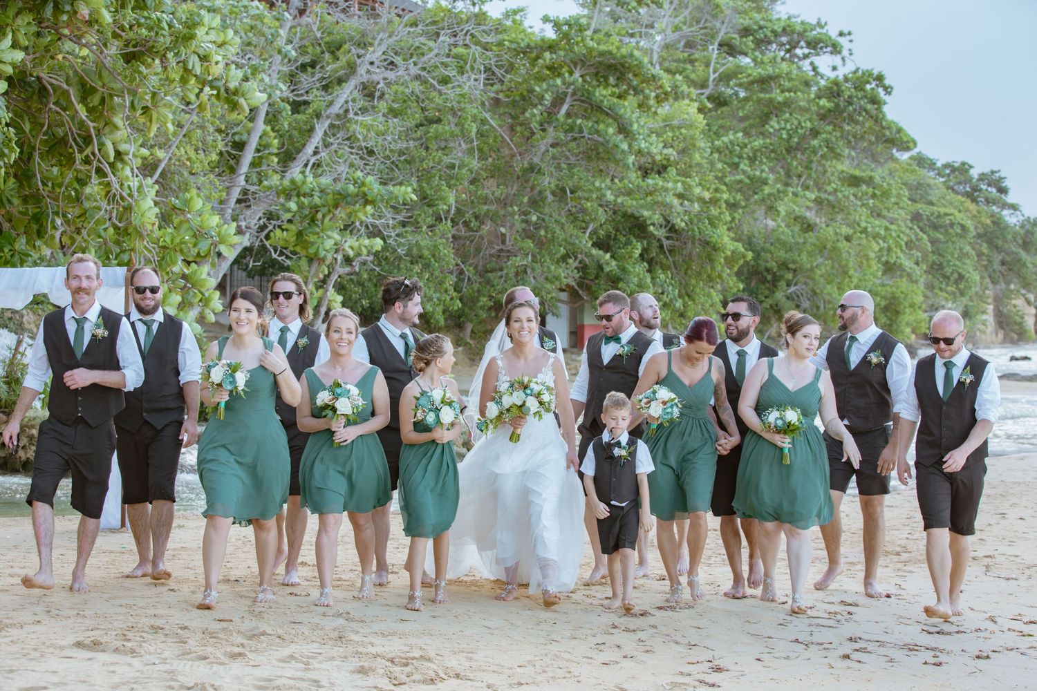 Wedding party in green and dark attire walking together on a sandy tropical beach.