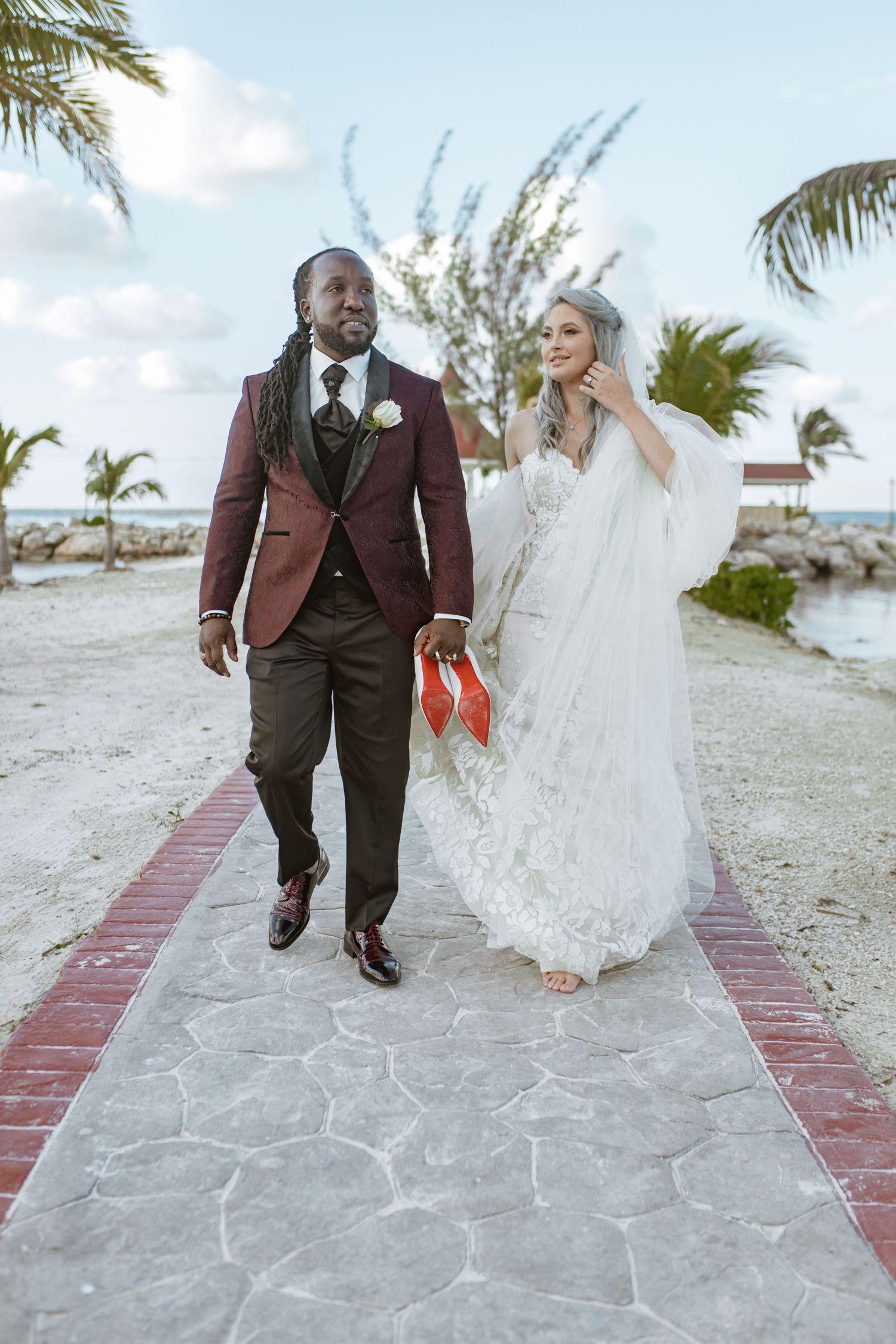 Newlywed couple walks along beachside pathway, bride in white lace gown holding red shoes.