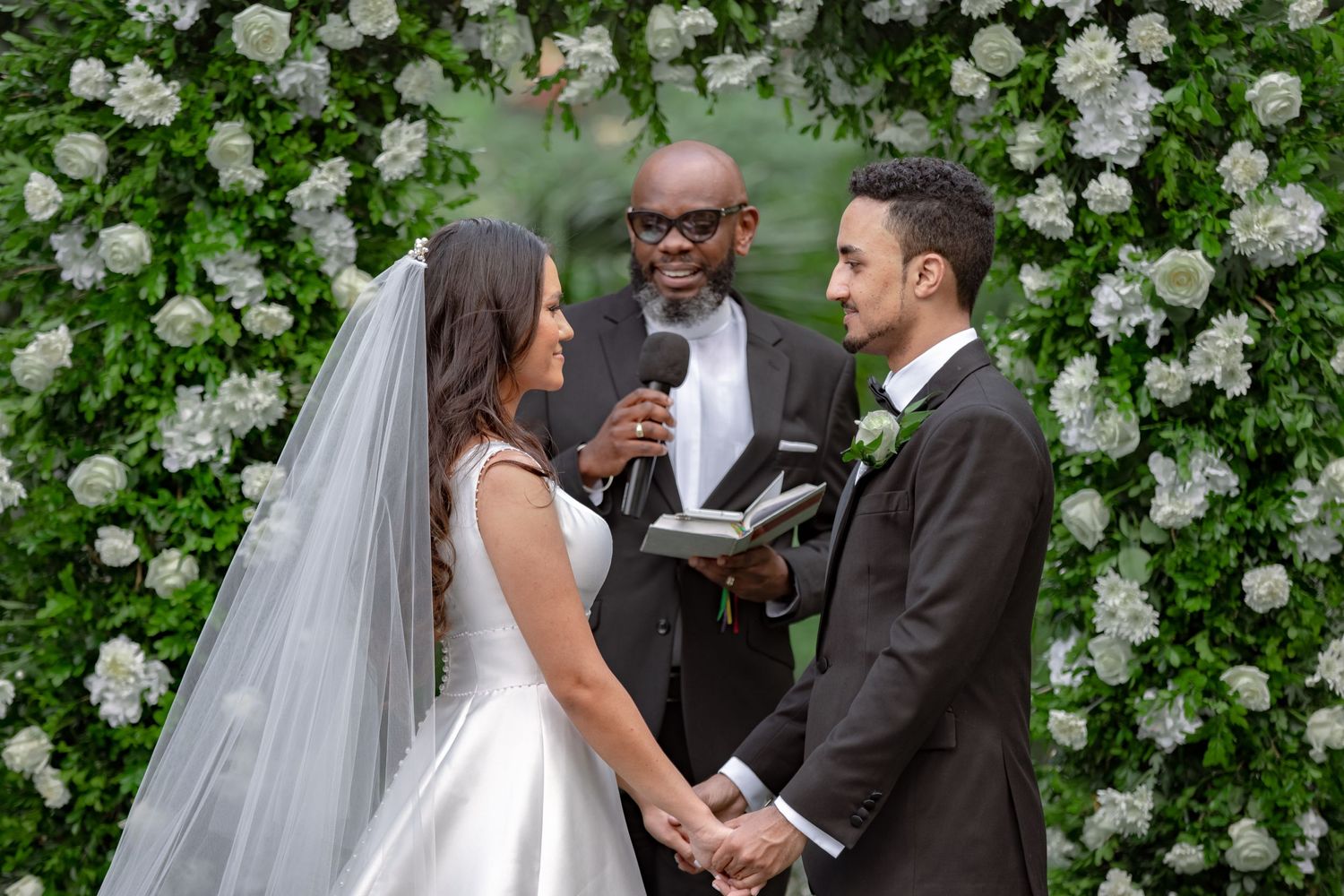 Bride and groom exchanging vows before officiant at white floral wedding altar.