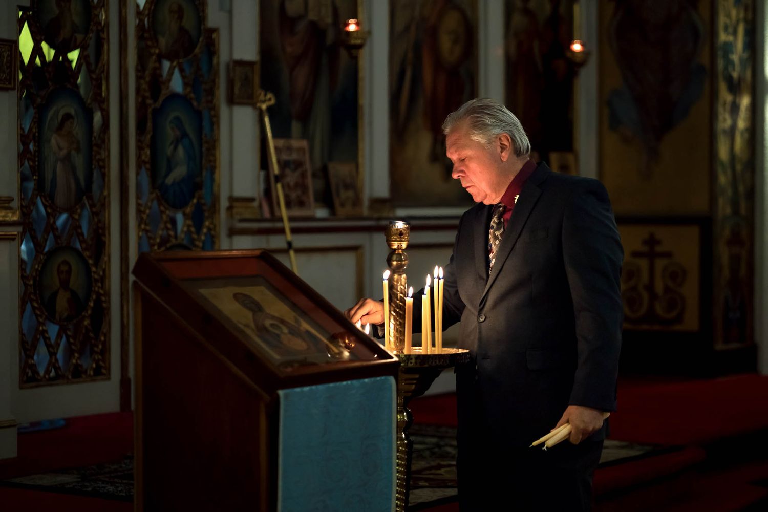 Parishioner standing in a dimly lit Eastern Orthodox sanctuary lighting a candle in prayer.
