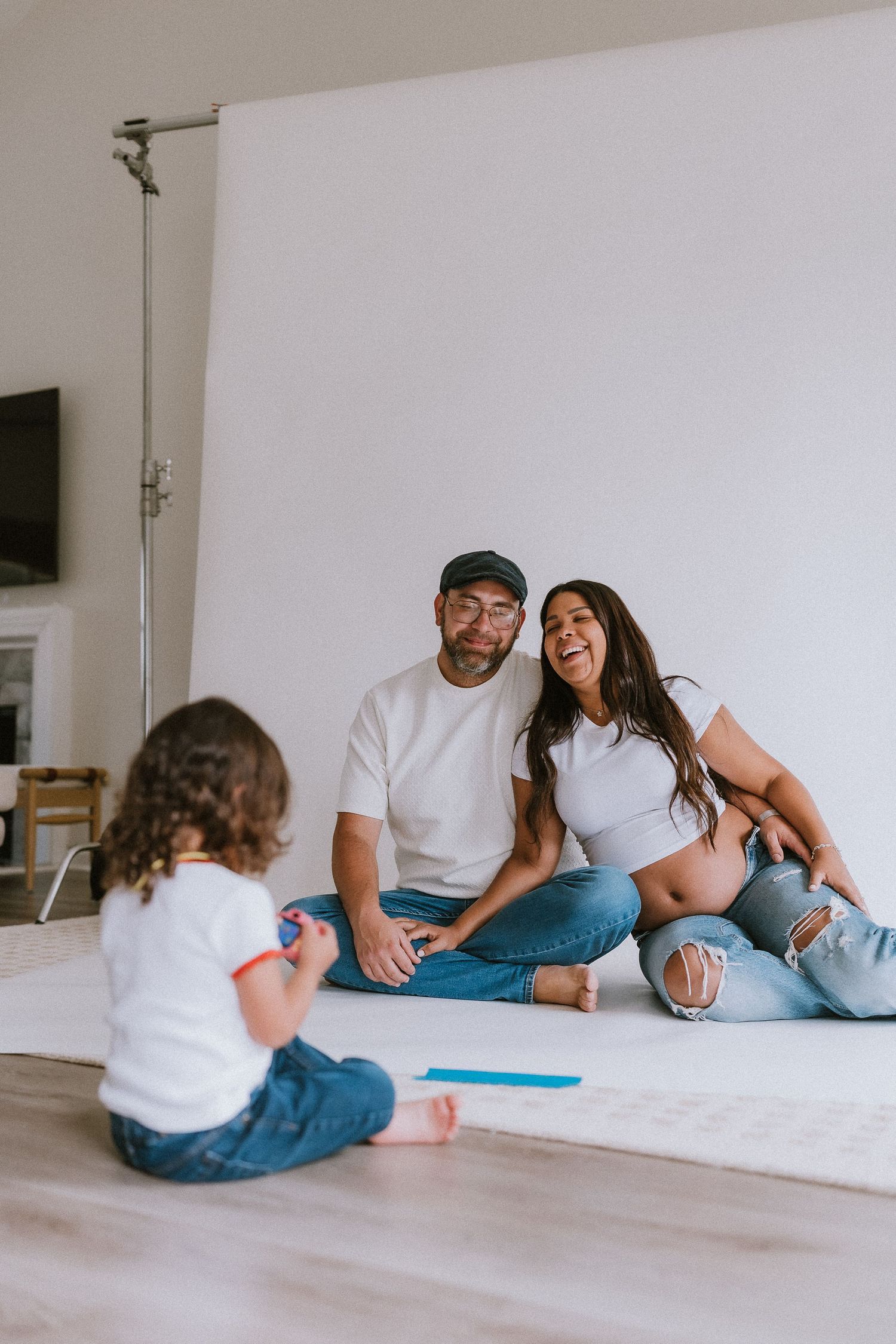 Pregnant woman and man in white shirts and jeans sit on floor laughing while toddler plays in front of white backdrop.