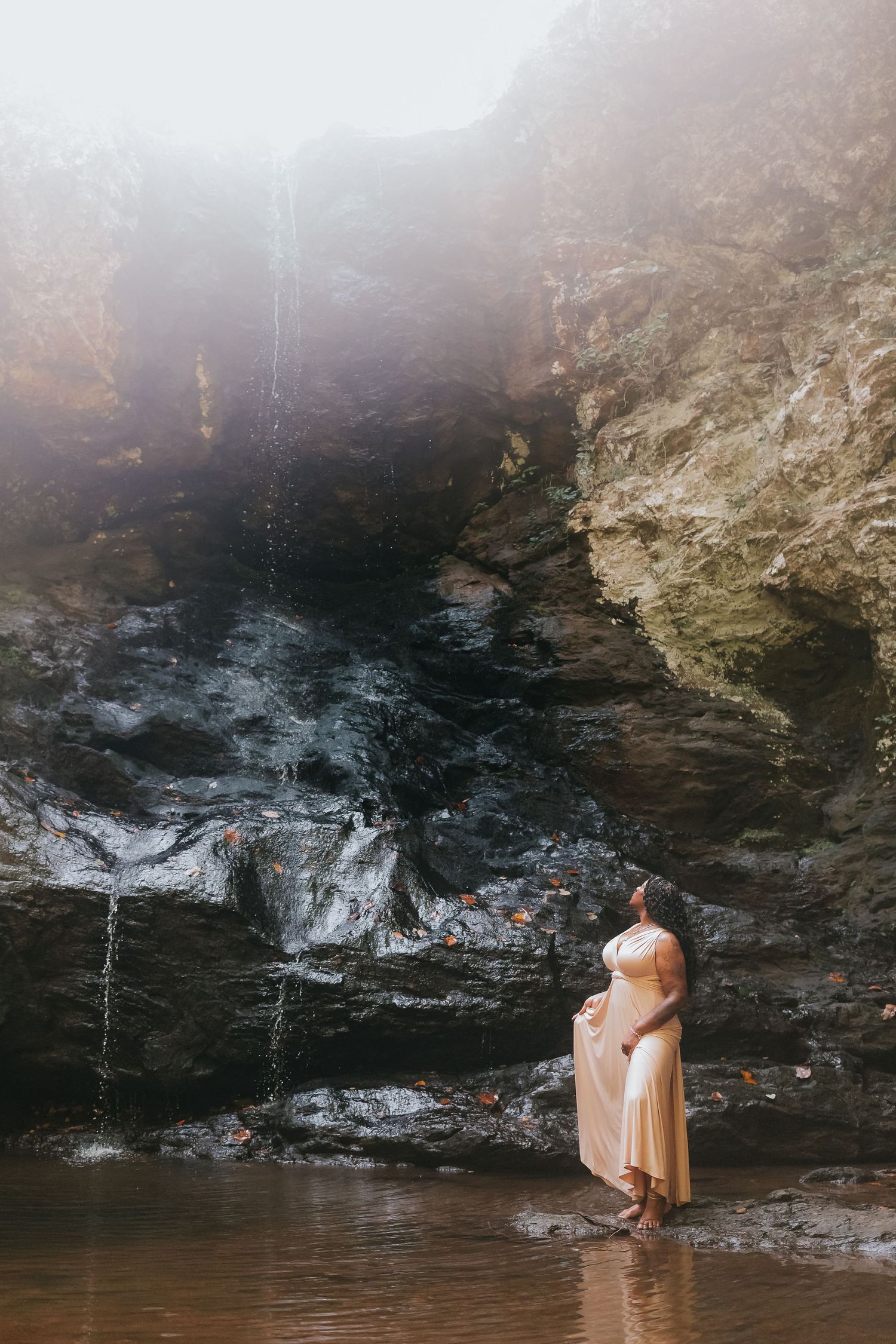 Woman in cream dress standing near rocky waterfall cave, bathed in soft misty light.