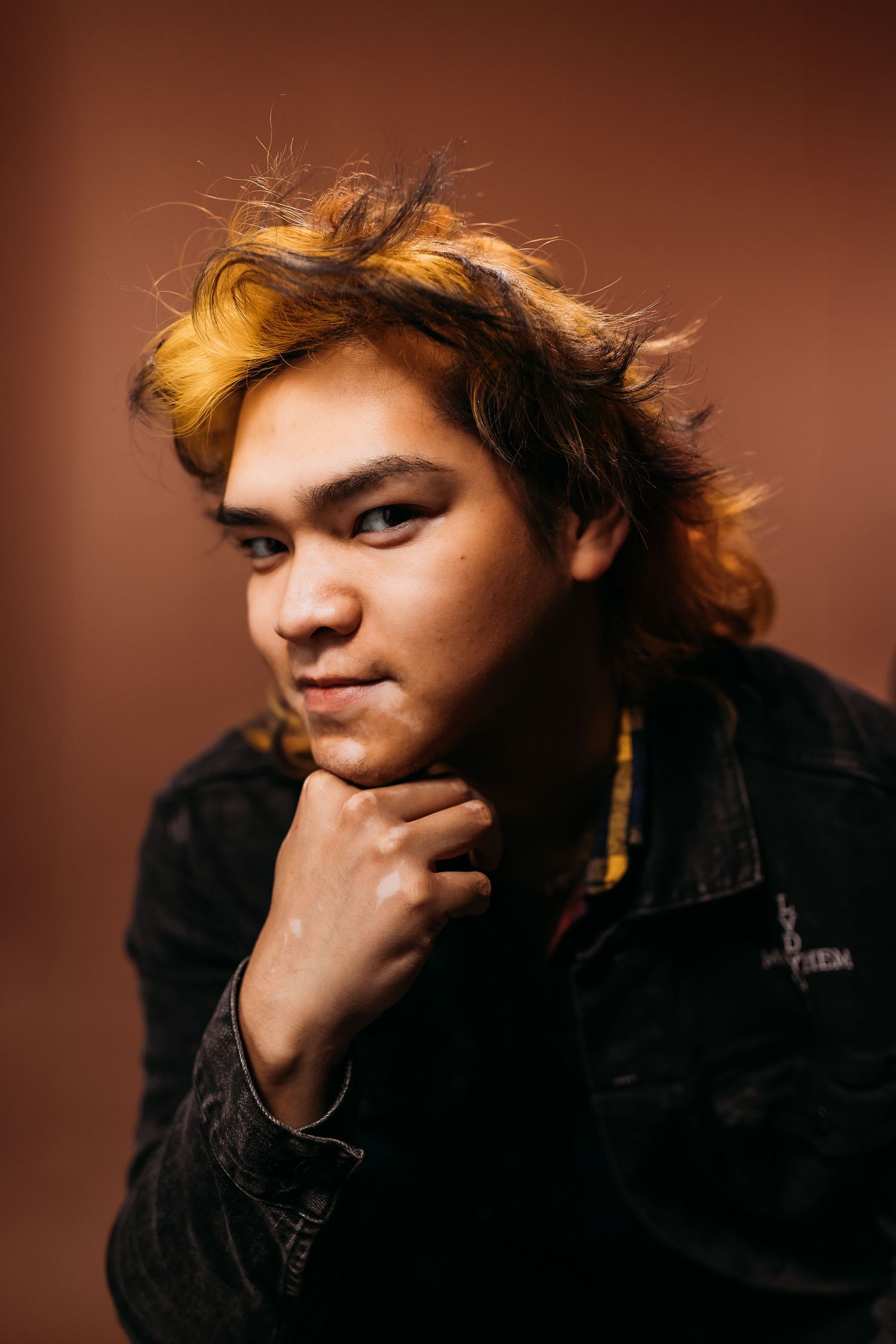 Young man with dyed blonde hair in black jacket posing thoughtfully against brown background.