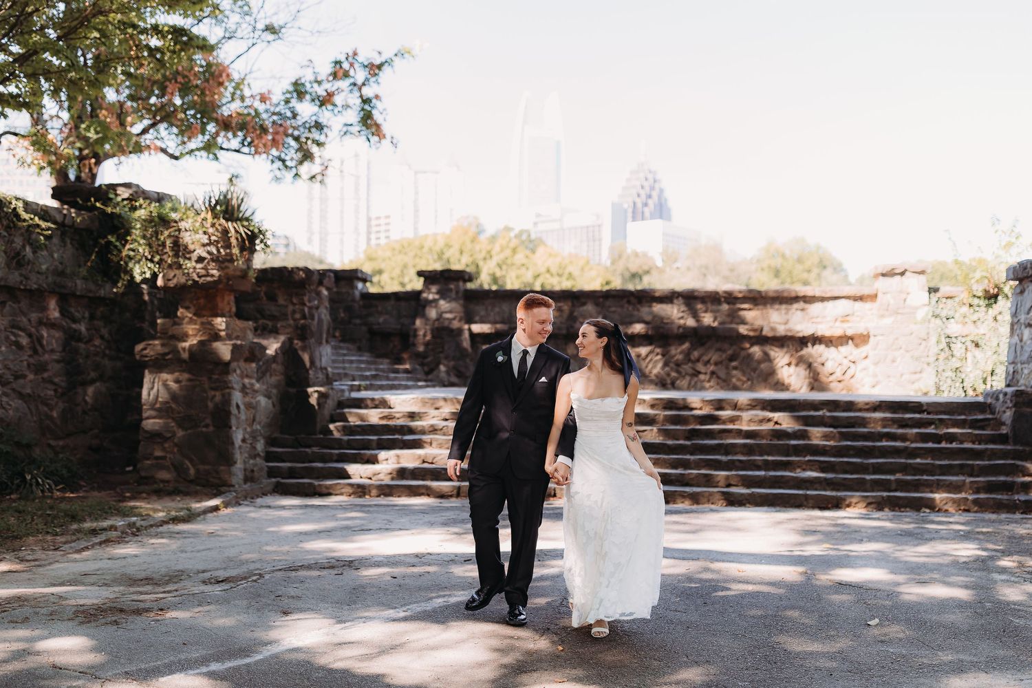 Bride in white gown and groom in dark suit walk hand in hand past stone steps and ruins with a hazy city skyline behind them.