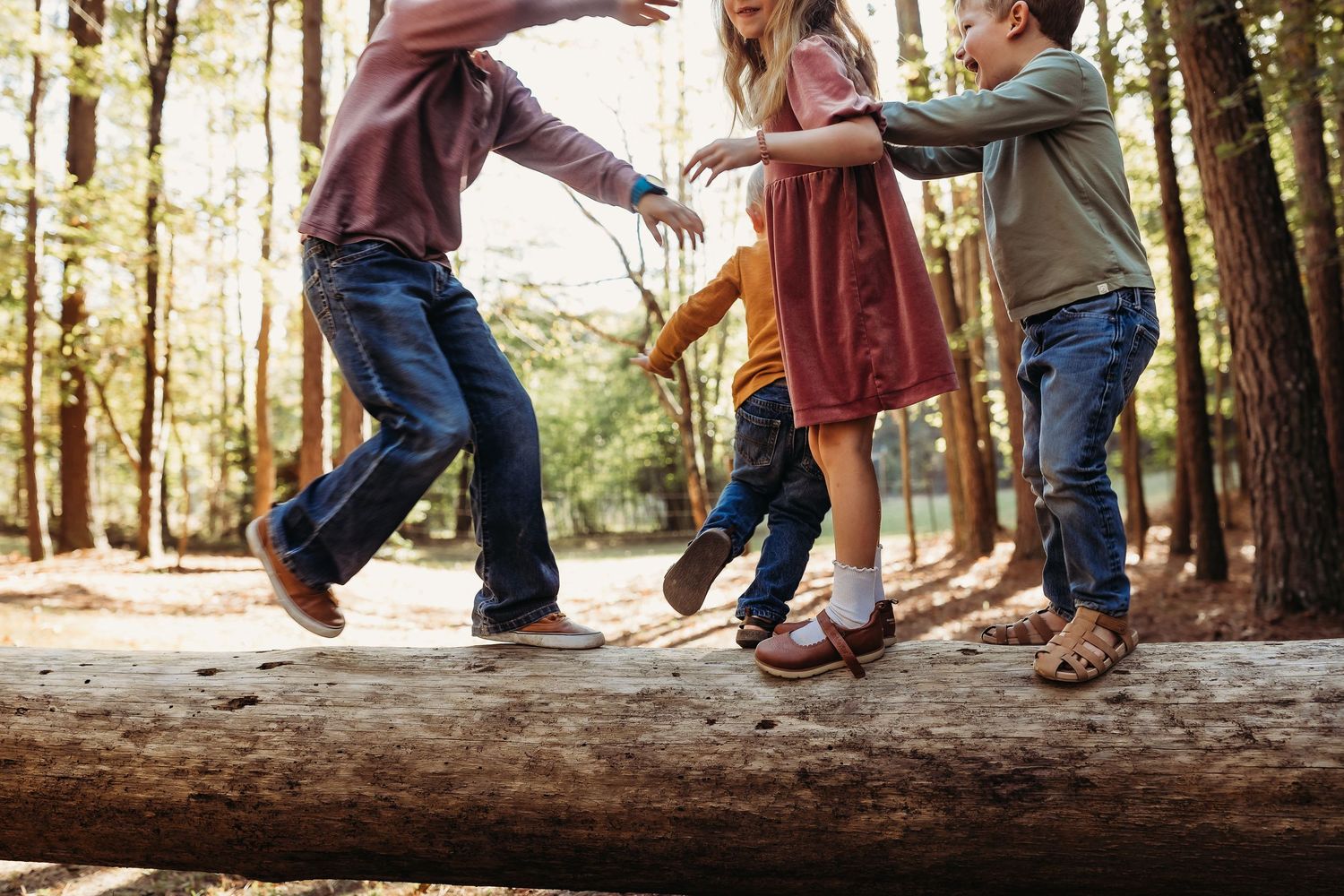 Children balancing on a fallen log in a sunlit forest, playing outdoors together.