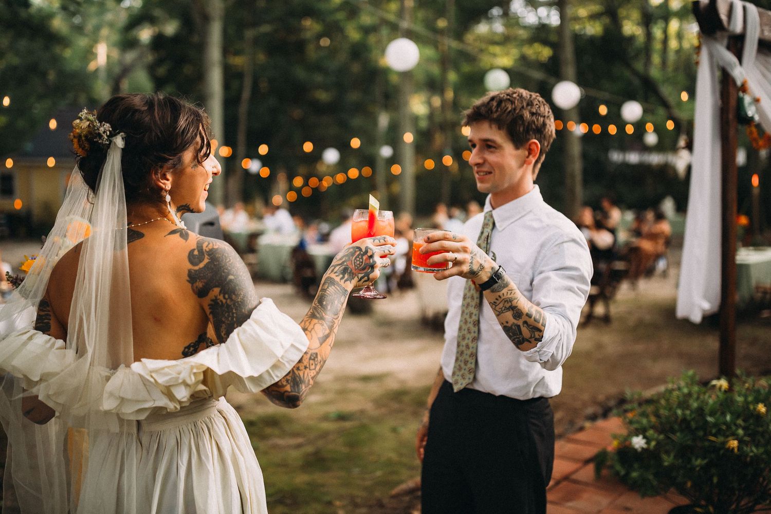 Tattooed bride and groom toasting with drinks at an outdoor woodland wedding reception.
