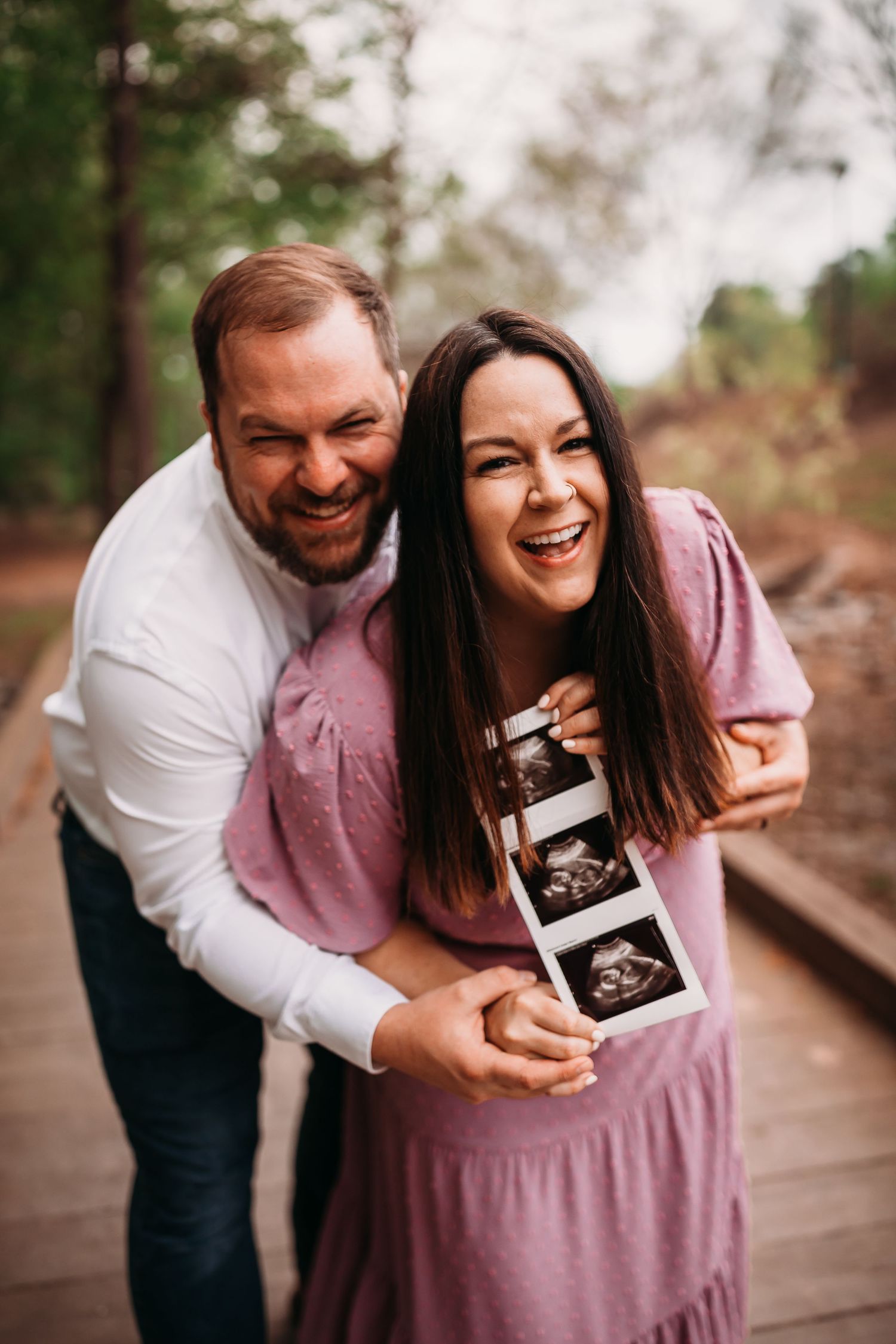 Happy couple holding ultrasound photos on a wooden bridge, announcing their pregnancy outdoors.