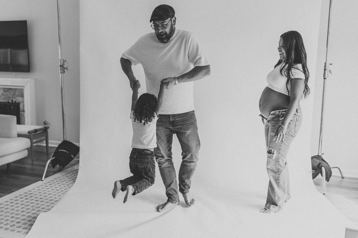 Black and white maternity photo of pregnant woman watching man swing toddler in a photography studio. Acworth, GA