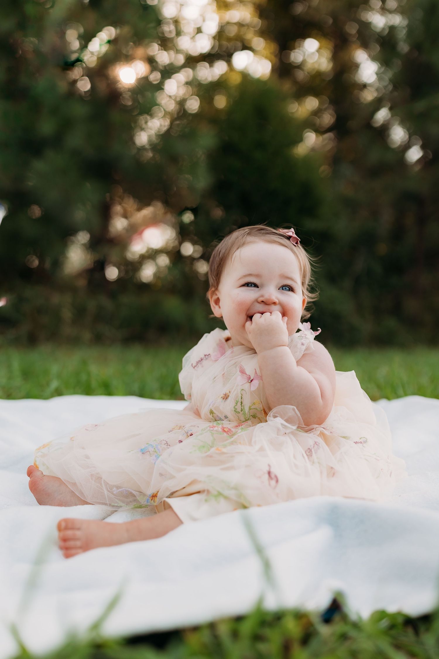 A happy baby girl in a floral dress sits on a white blanket outdoors, smiling with her hand near her mouth.