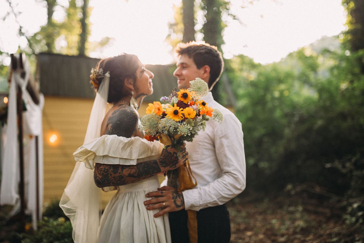 Tattooed bride and groom sharing a tender moment holding sunflower bouquet in forest setting.