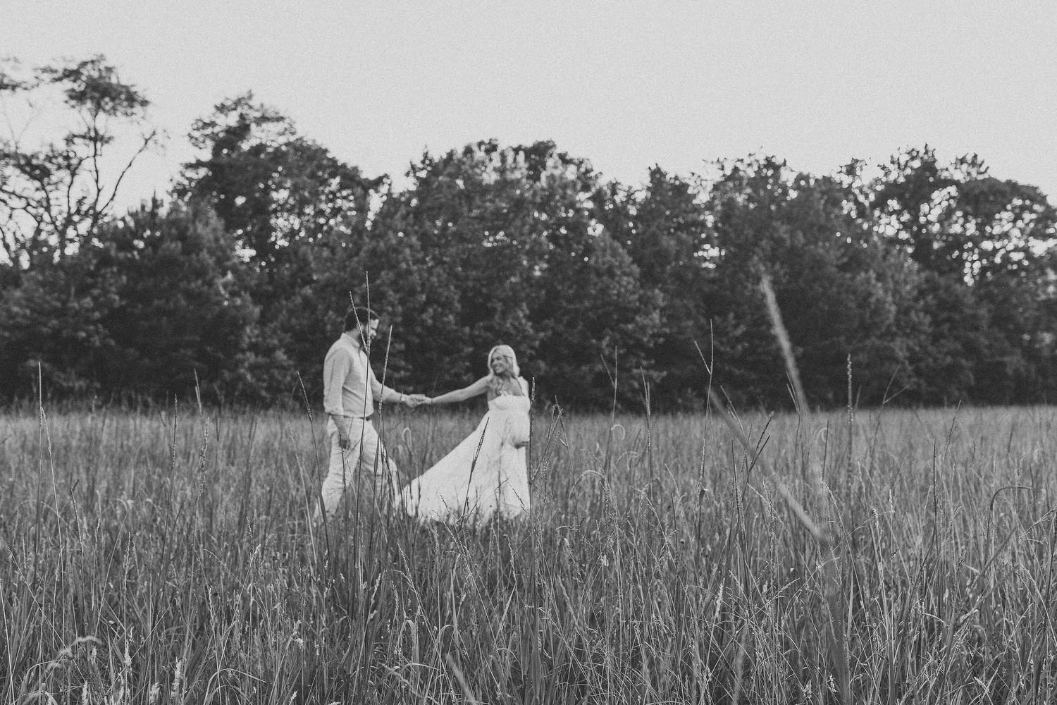 Black and white photo of a couple dancing in a field of tall grass with trees in the background.