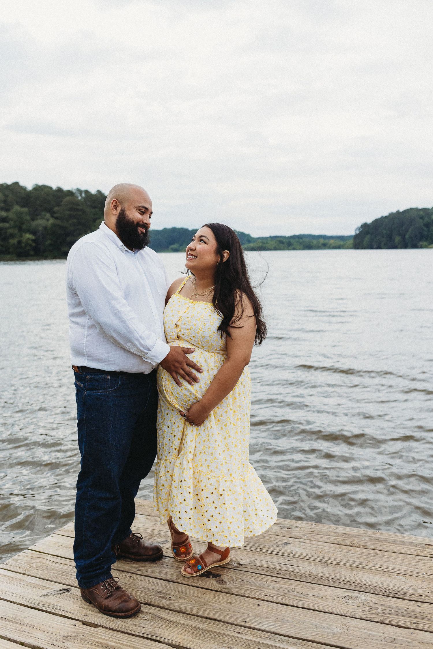 Pregnant couple sharing a tender moment on a wooden dock by a lake, woman in yellow dress cradling her baby bump.