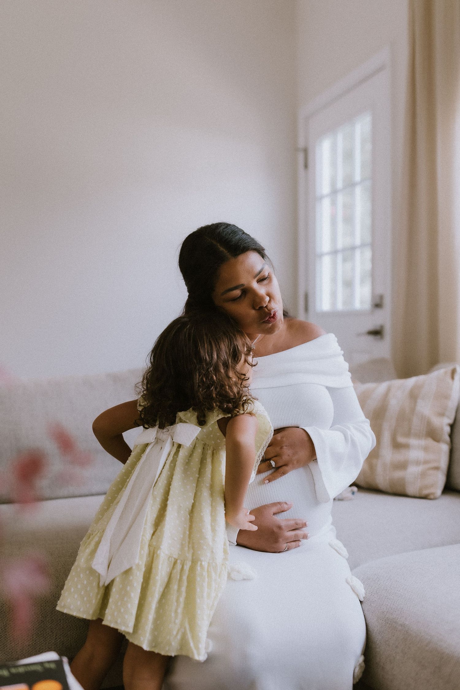 Pregnant woman in white dress hugging young girl in yellow dress on a couch in a bright living room.