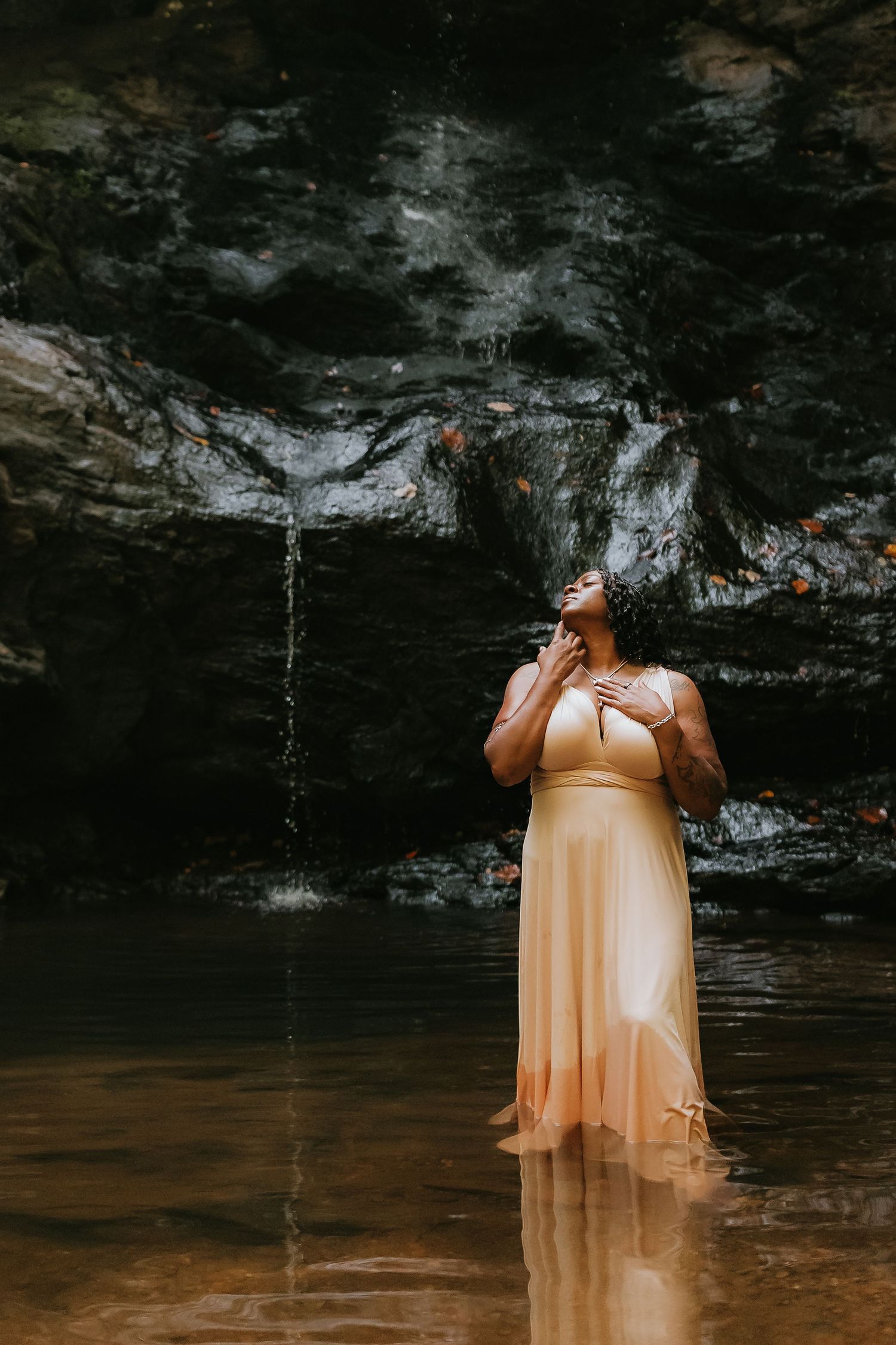 Woman in peach gown standing in shallow water before a rocky waterfall, gazing upward in serene contemplation.