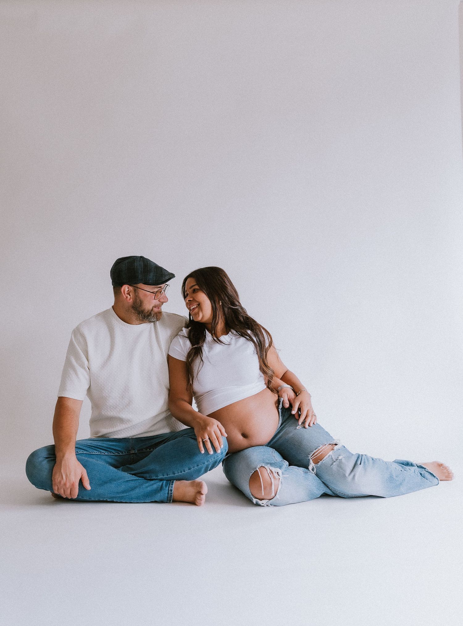 Couple sitting on floor smiling at each other, pregnant woman in white crop top and ripped jeans, man in white tee and cap. Kennesaw, GA