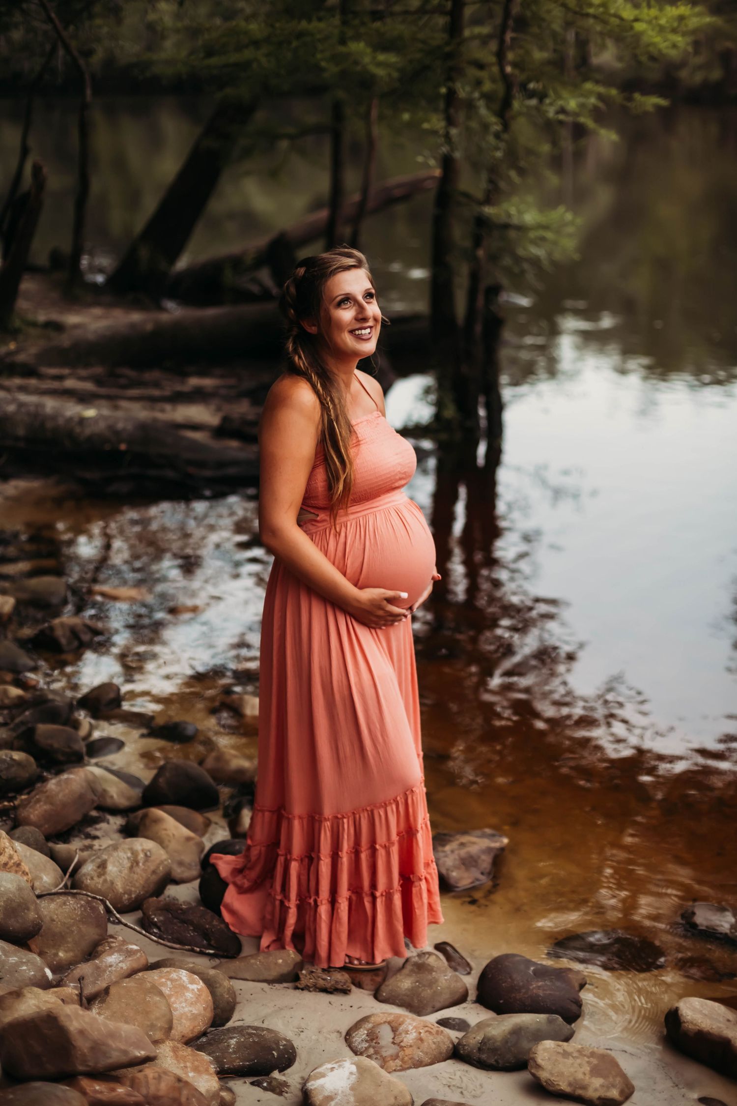 Smiling pregnant woman in coral maxi dress standing on rocky riverbank surrounded by lush green trees.