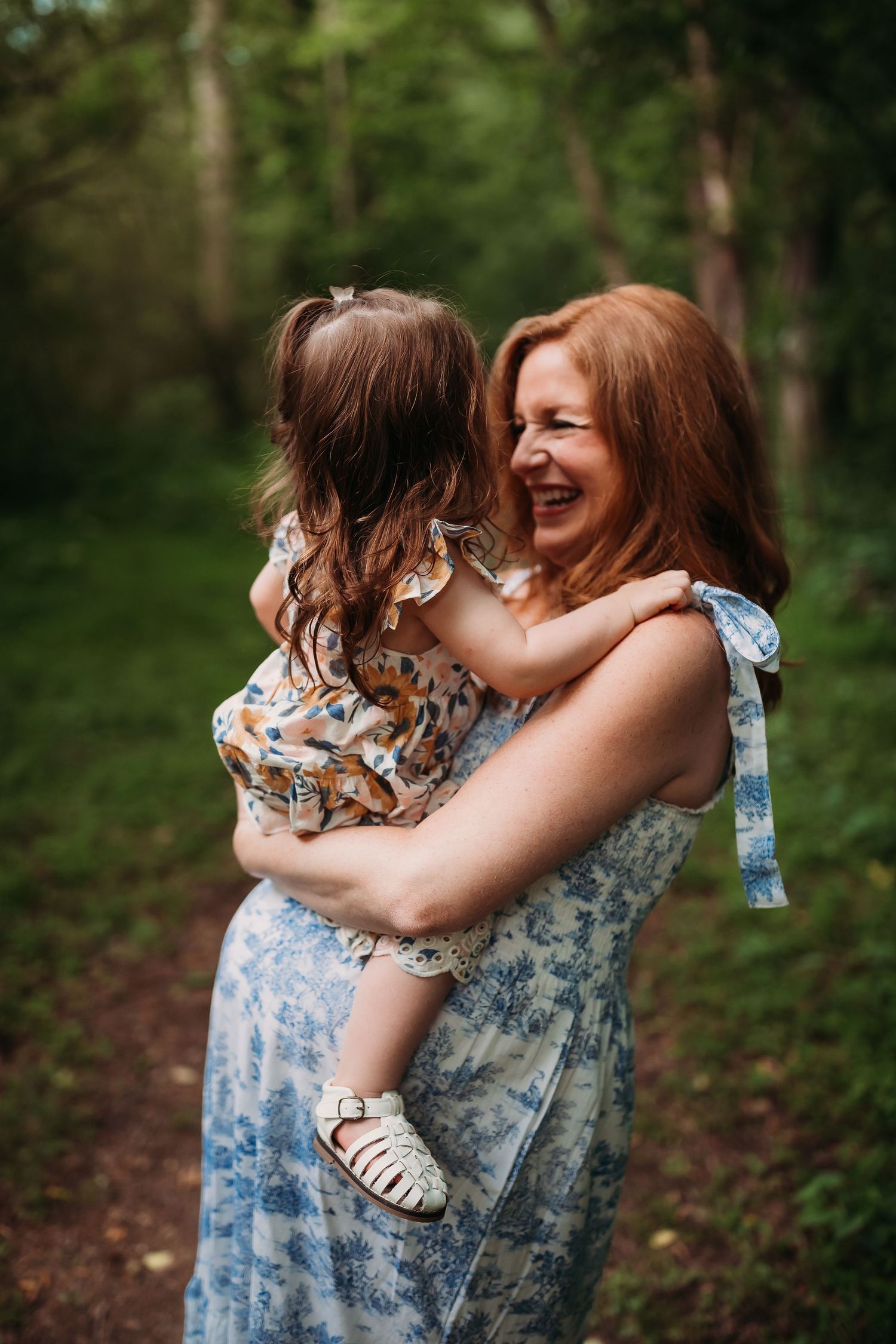 A laughing redhead woman in a floral dress holds a young girl in a woodland setting.
