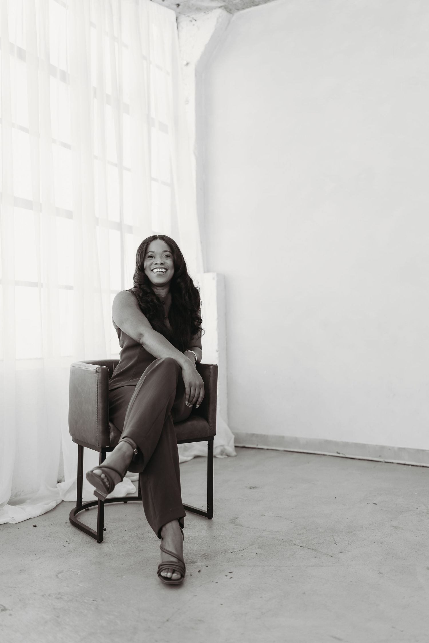 Black and white portrait of a smiling woman with long hair sitting relaxed in a chair by a bright window.