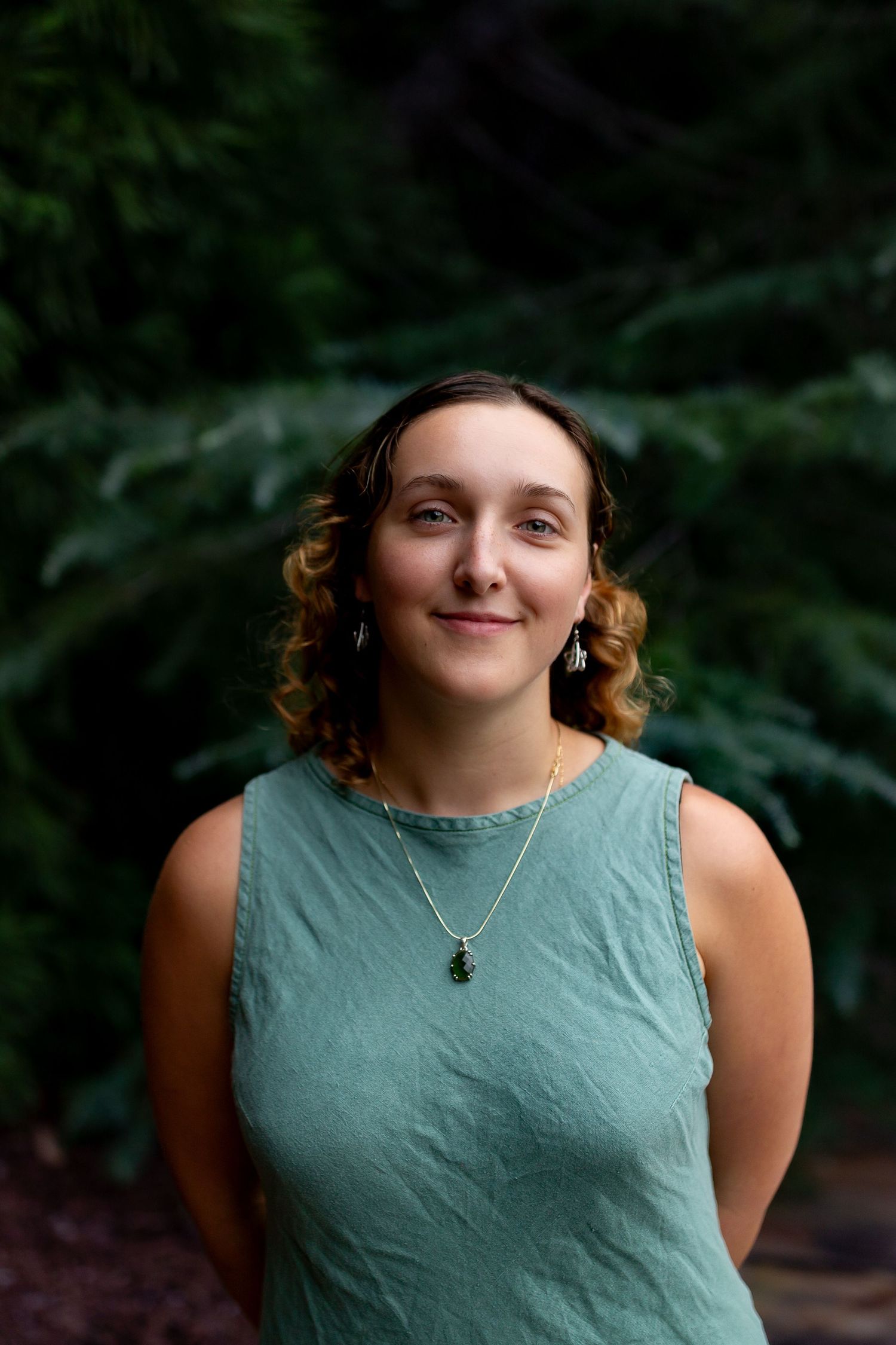 Young woman with curly hair smiling outdoors, wearing teal sleeveless dress and green pendant necklace.