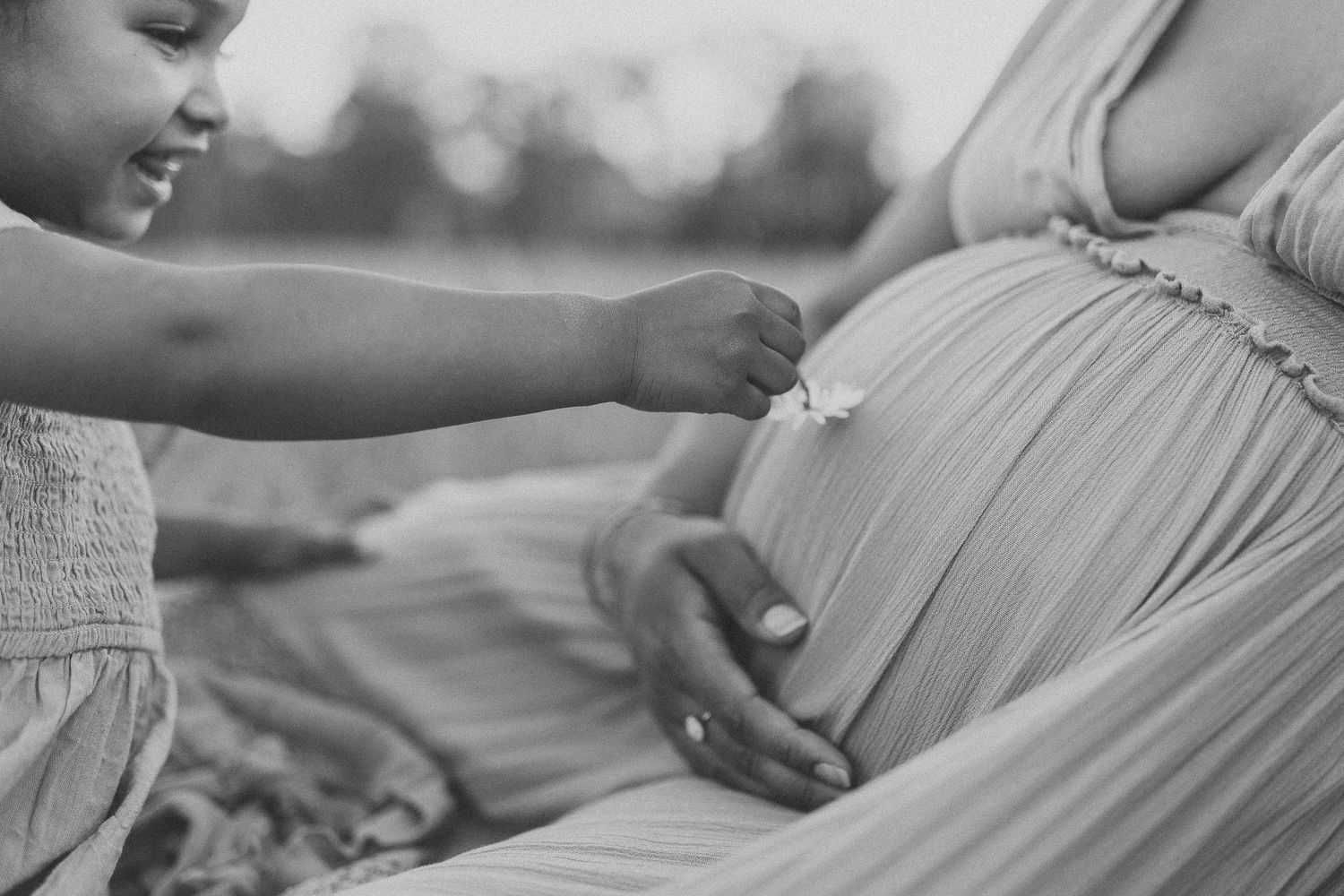 Black and white photo of a young girl placing a flower on her pregnant mother's baby bump outdoors.