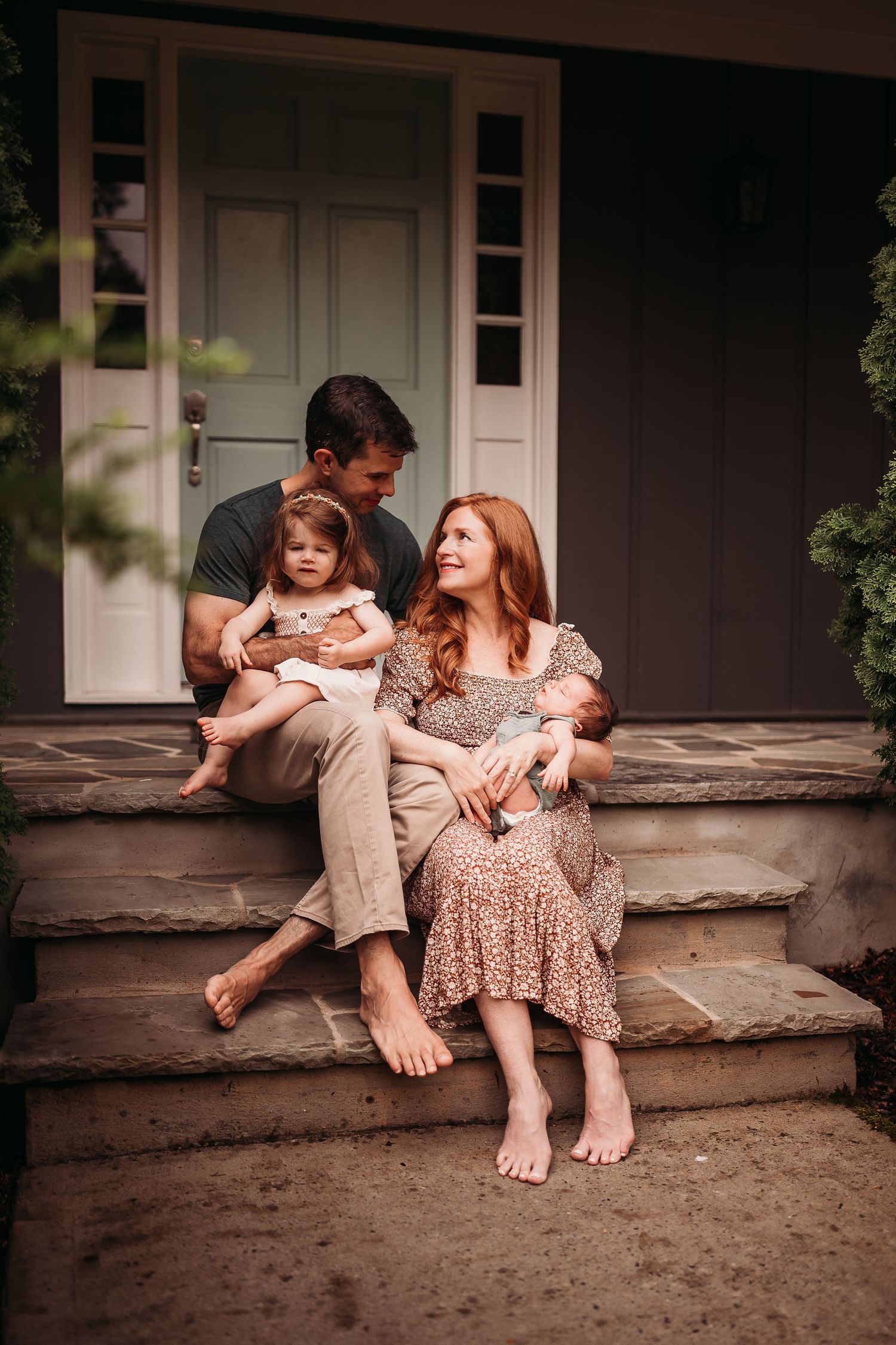 A happy family sits barefoot on stone porch steps, with dad holding toddler and mom cradling newborn baby.