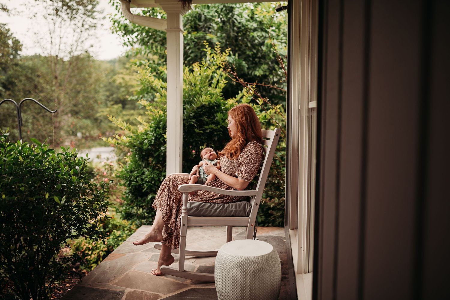A red-haired mother breastfeeding her newborn in a rocking chair on a serene porch surrounded by lush greenery.