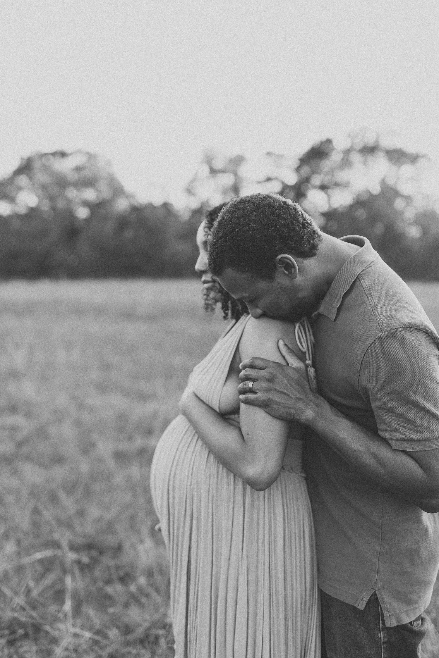 Black and white maternity photo of a couple embracing in a field, man kissing pregnant woman's shoulder tenderly.