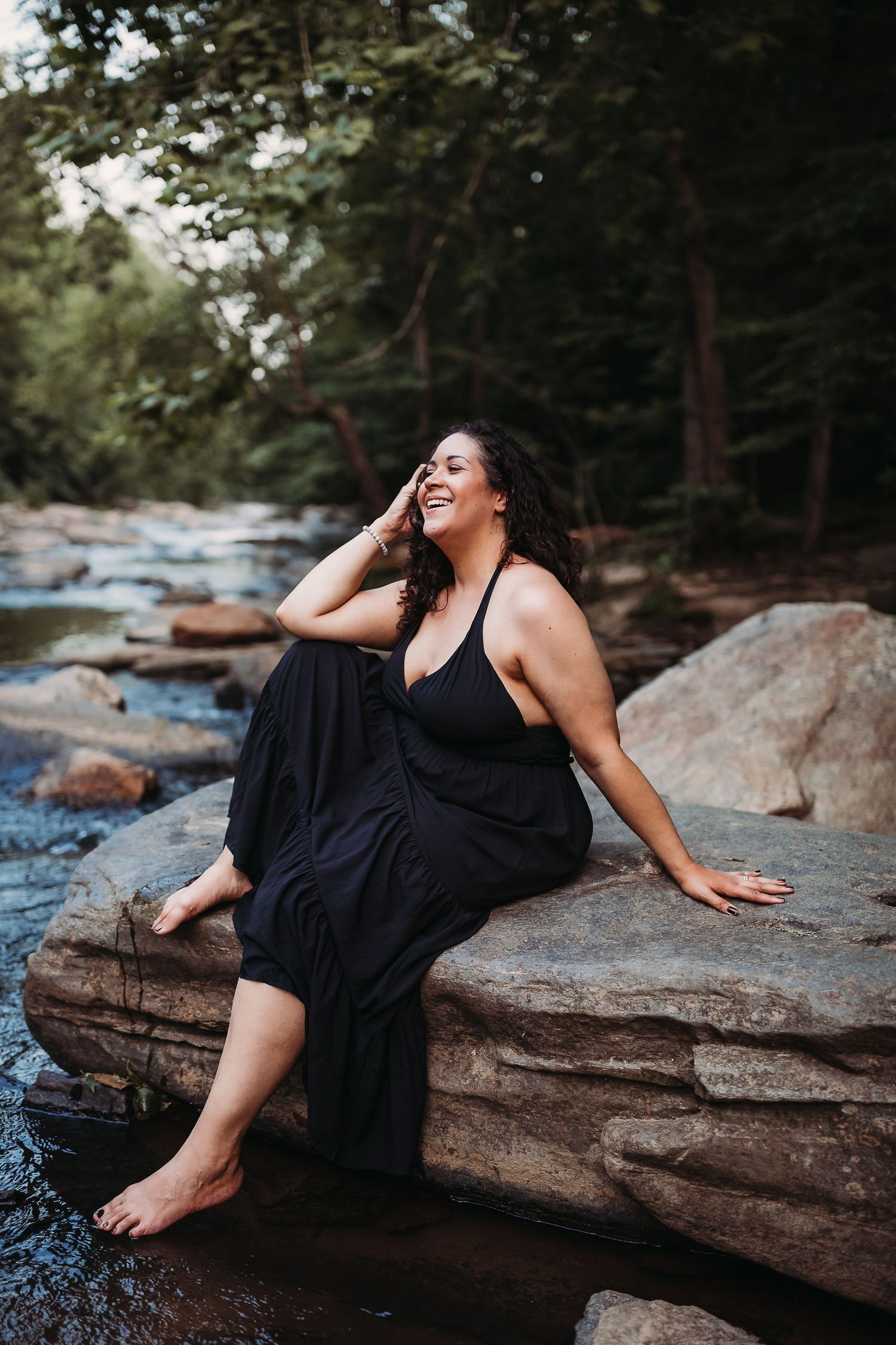 Woman in black dress sitting on rocks by forest stream, laughing with hand in hair.