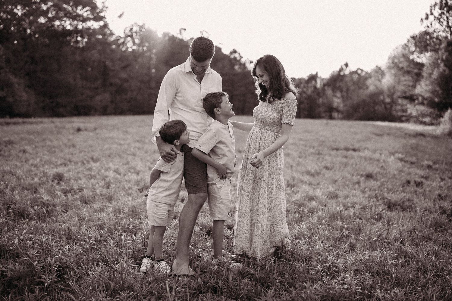 Black and white family portrait of parents with two children embracing in an open field at sunset.