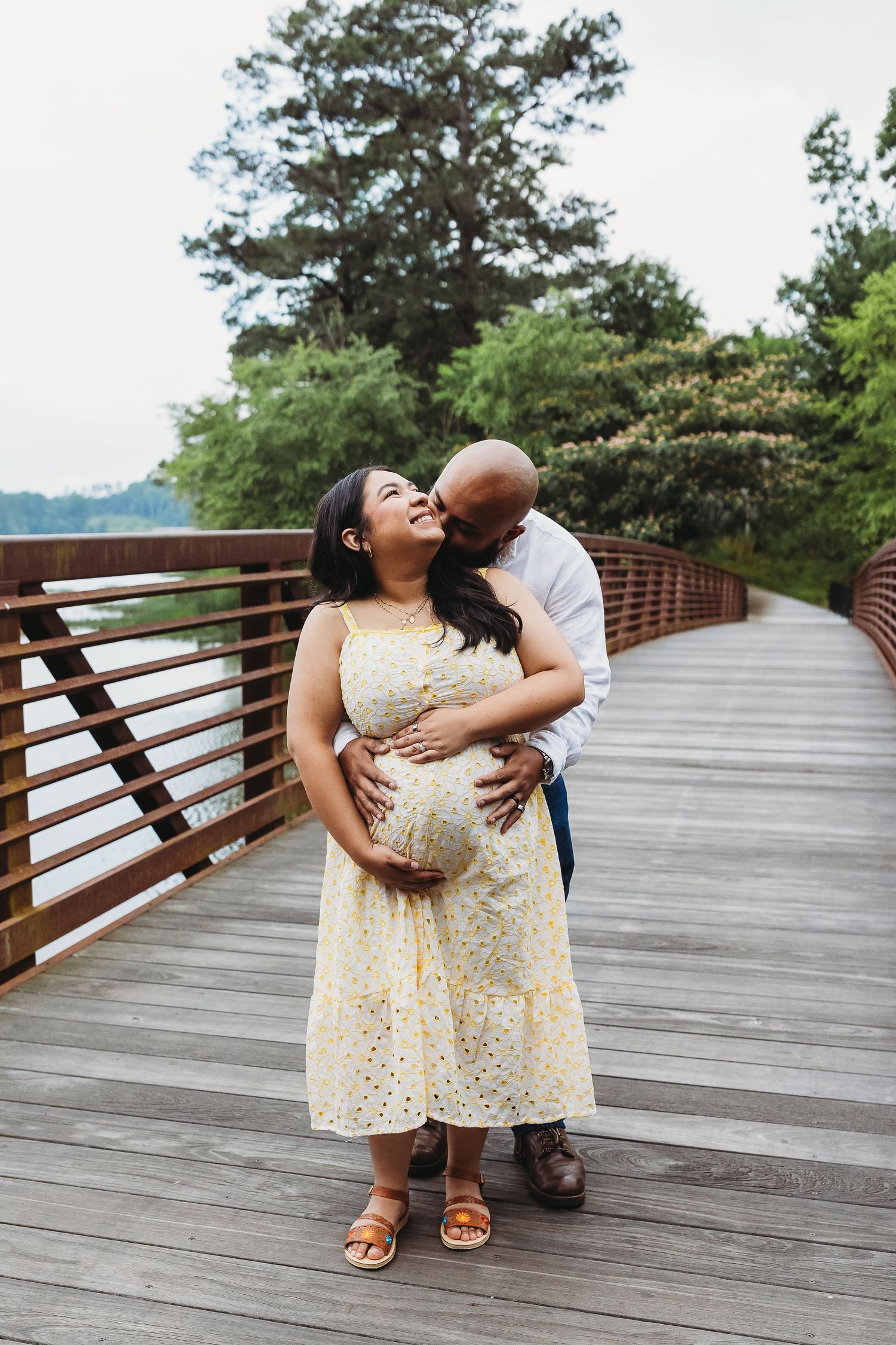 Pregnant couple embracing on wooden bridge, man kissing woman's cheek while both hold her baby bump, surrounded by lush greenery.