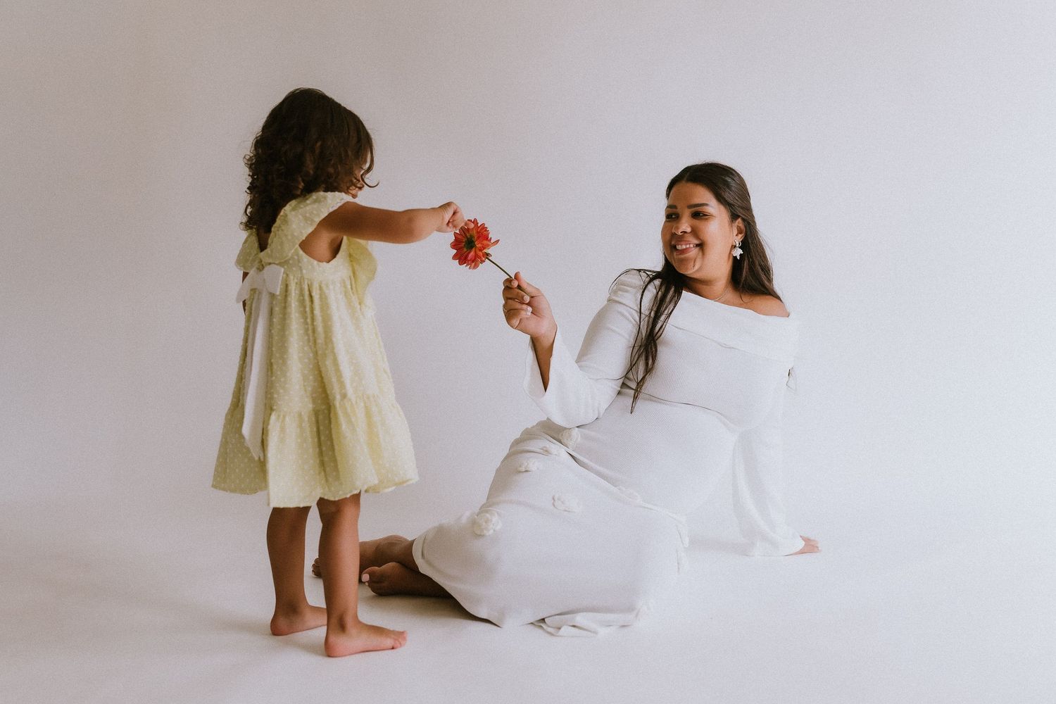 Pregnant woman in white dress sitting on floor receiving flower from young girl in yellow dress. Kennesaw, GA