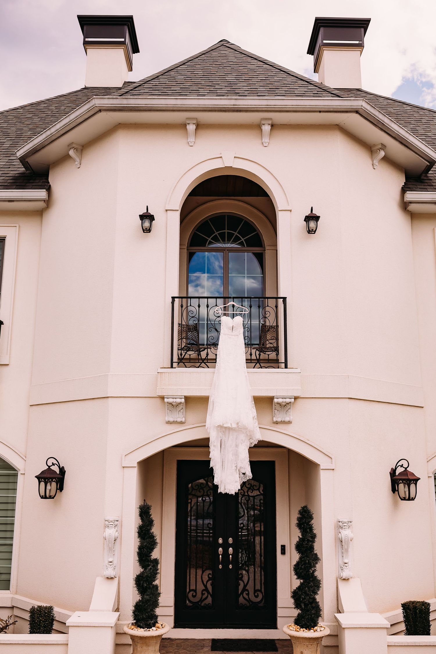 Elegant white wedding dress hanging from a balcony railing on a grand French-style estate entrance.