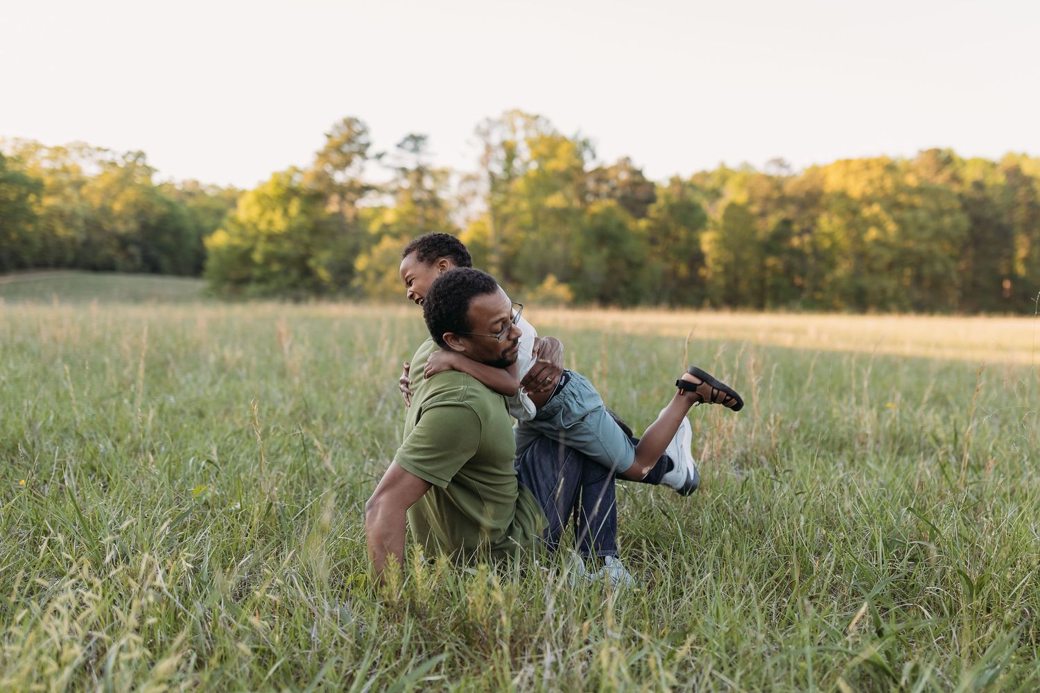 A couple playfully embraces in a lush green field at sunset, with the man lifting the woman joyfully.