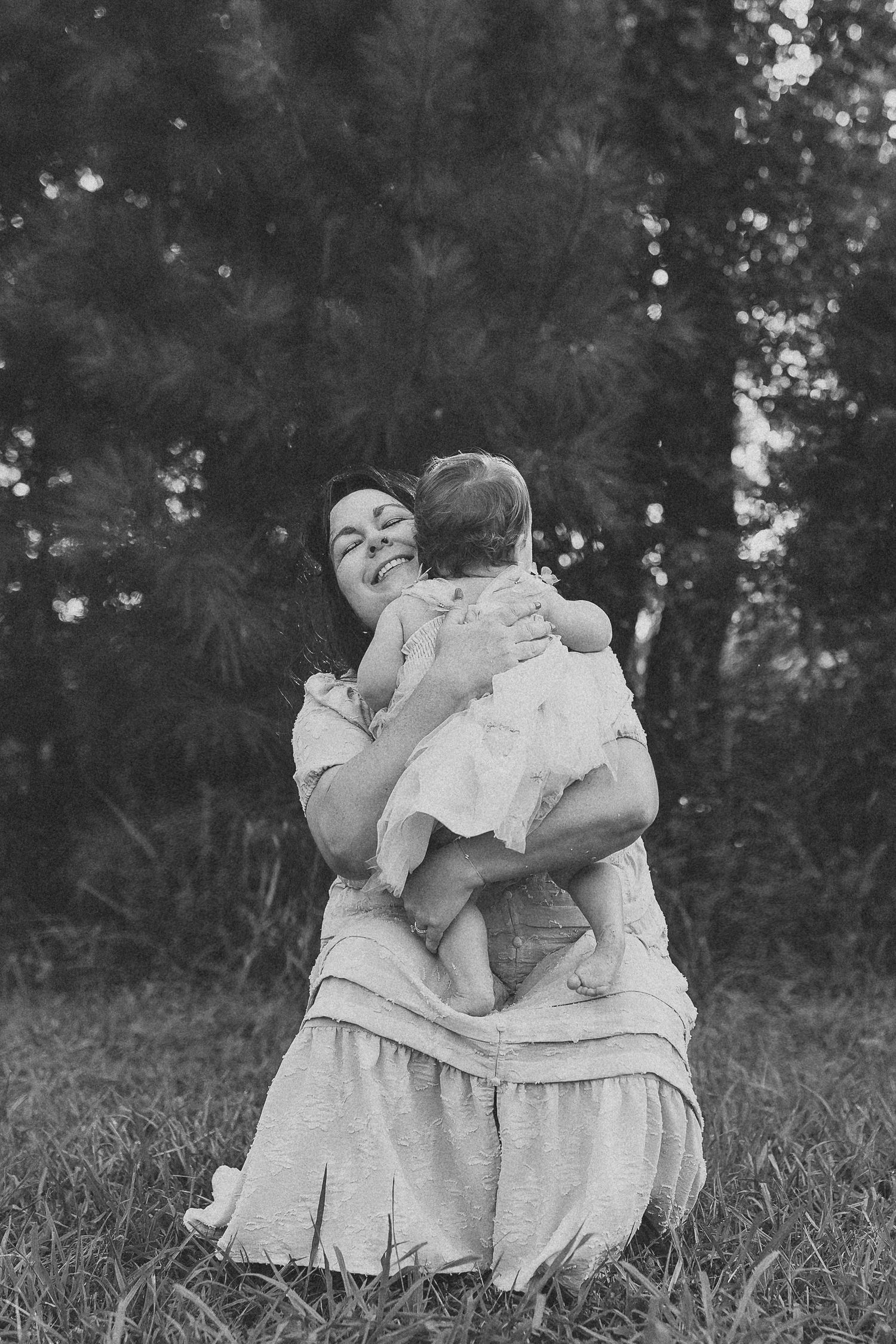 Black and white photo of a mother holding her baby outdoors, smiling joyfully while sitting on grass near trees.