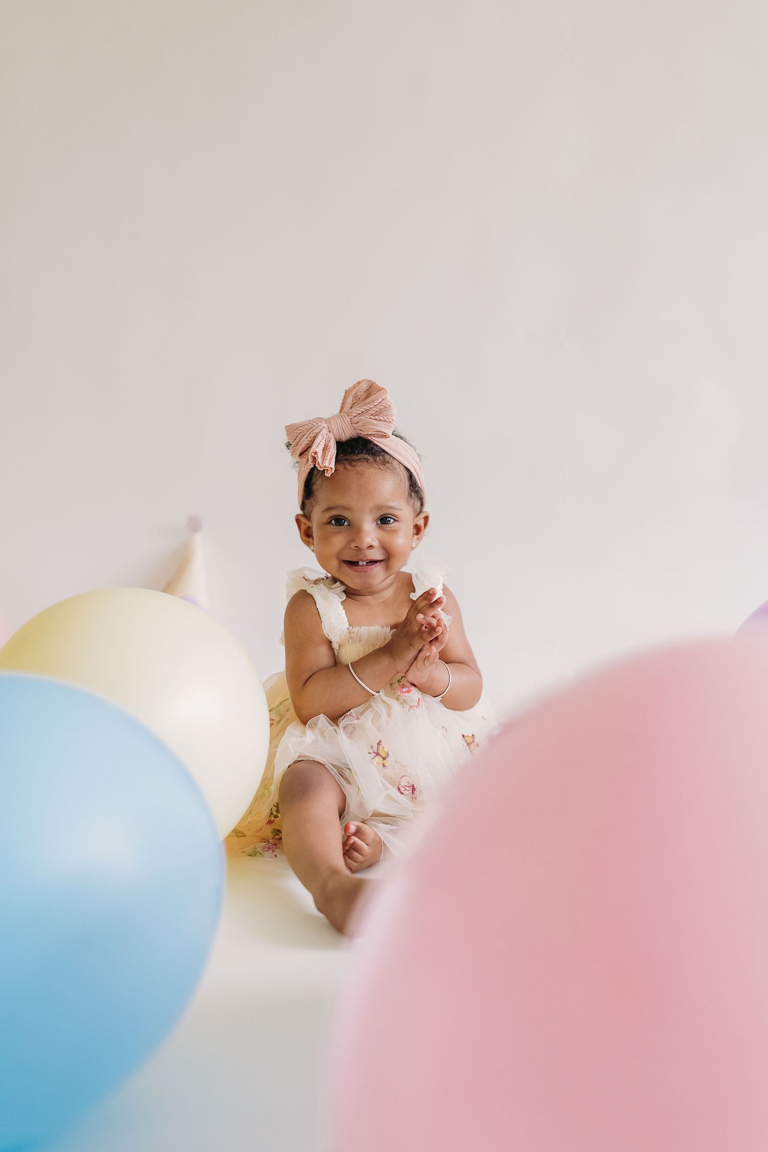 Smiling baby girl in white dress and pink bow headband sitting among colorful pastel balloons at birthday celebration.