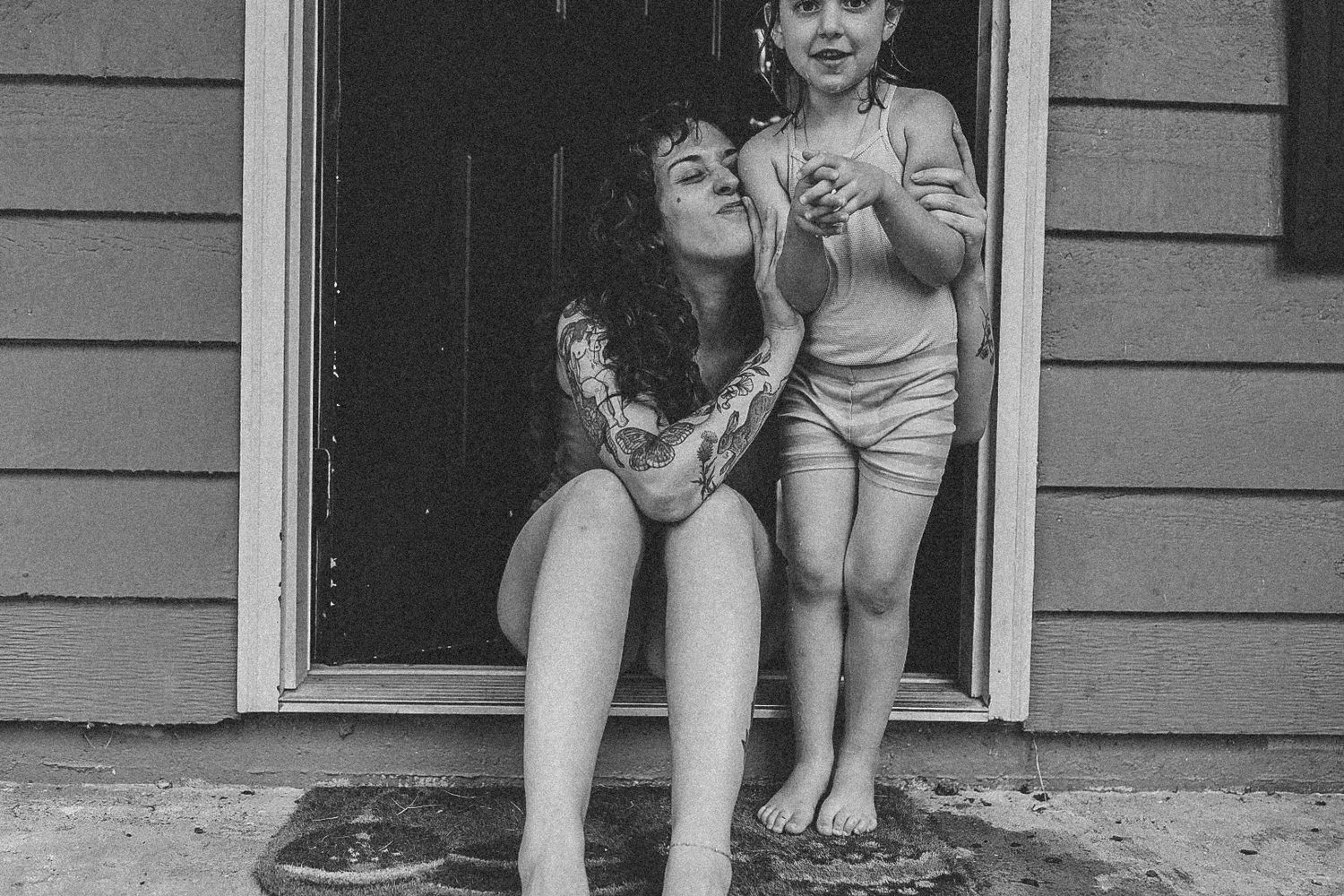 Black and white photo of tattooed woman and young child sitting in doorway during rain.