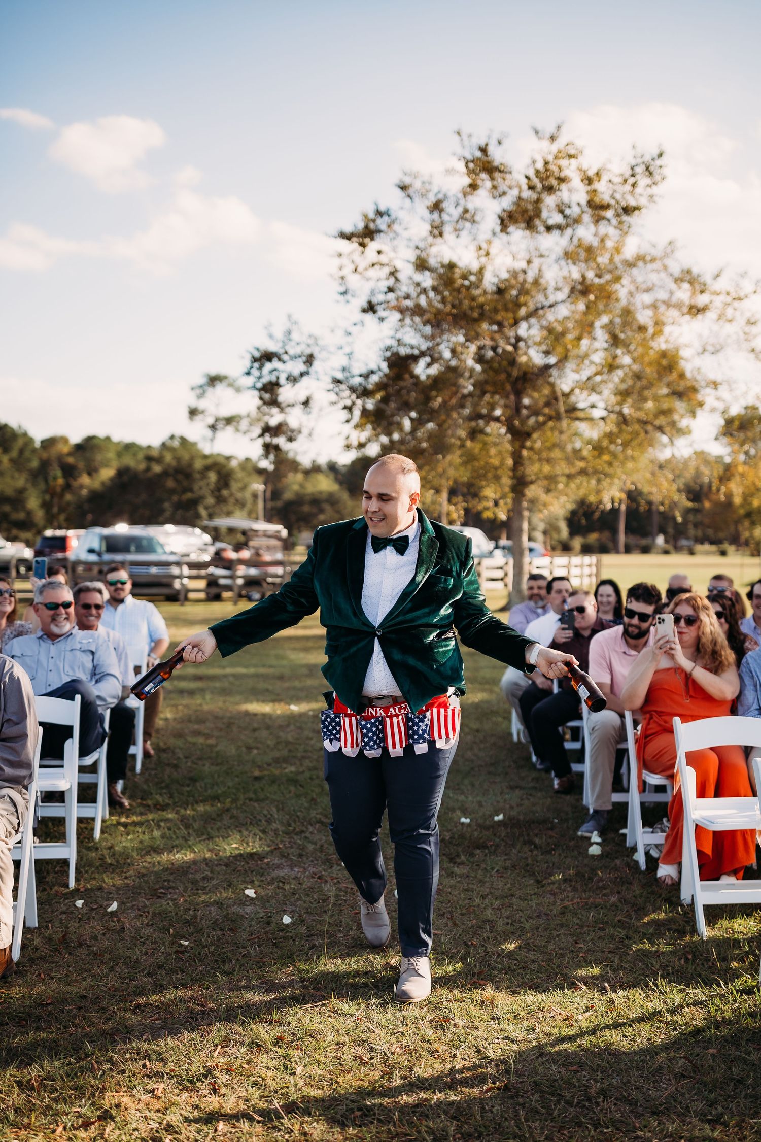 Energetic usher in green velvet jacket with patriotic accessories dances down outdoor wedding ceremony aisle.
