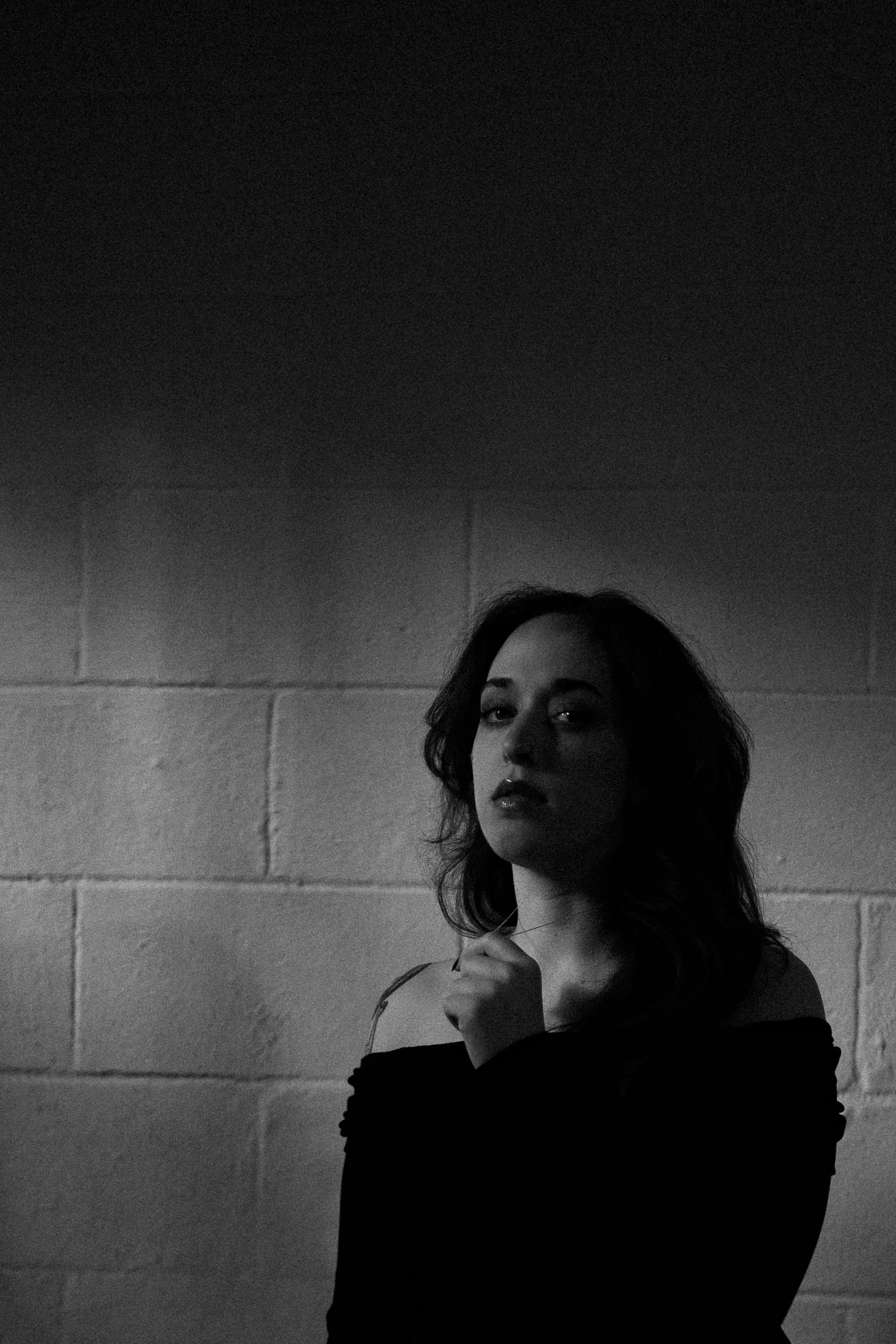 Dramatic black and white portrait of a young woman standing against a brick wall in dim lighting.