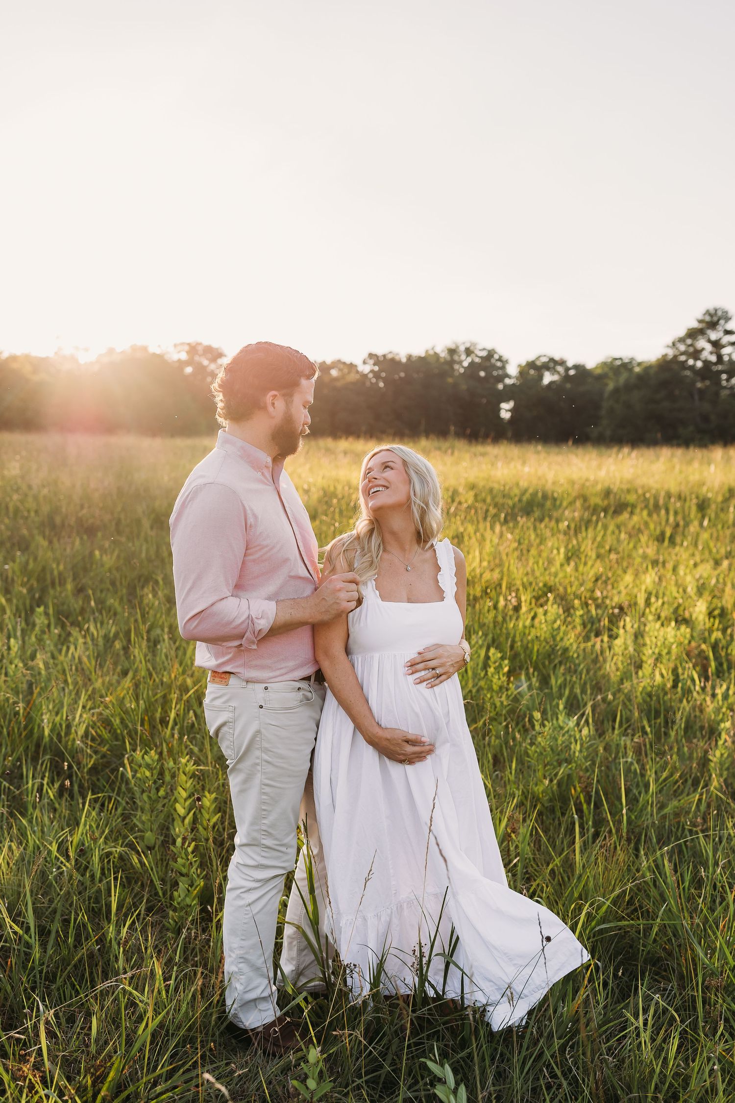 Pregnant woman in white dress laughing with partner in a sunlit green meadow during a golden hour maternity photoshoot.