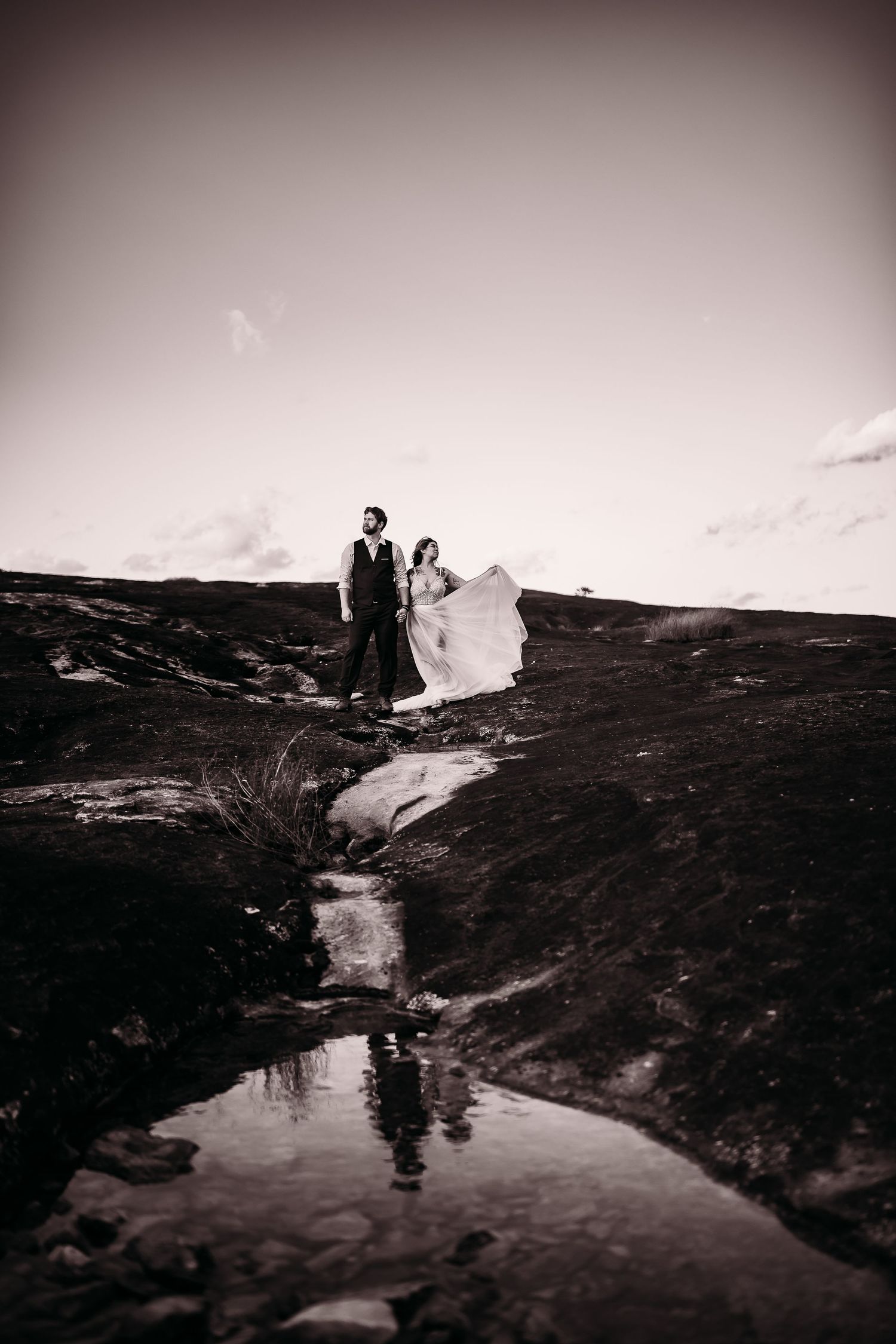 A black and white photo of a bride and groom standing on rocky landscape with a puddle reflection below.