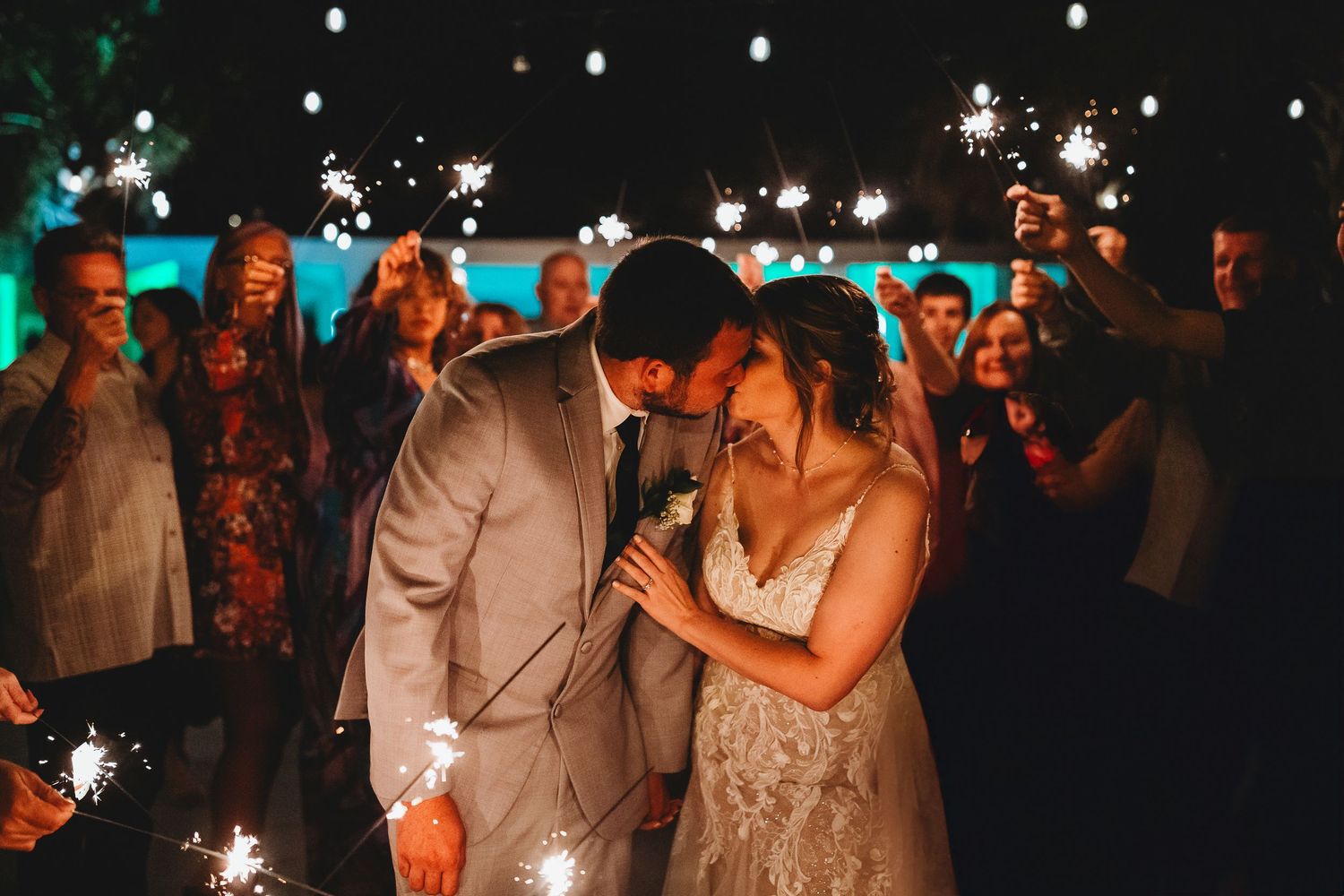 Bride and groom share a kiss surrounded by guests holding sparkling sparklers at their evening wedding reception.
