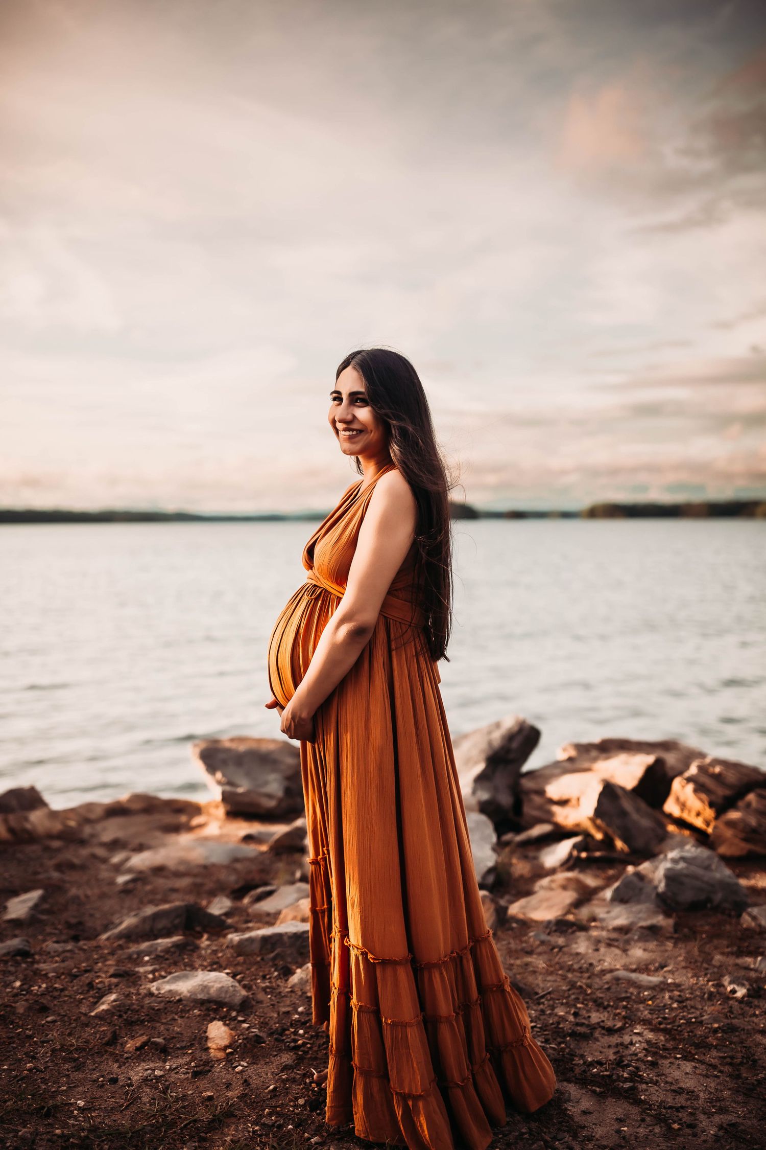 Smiling pregnant woman in rust orange maxi dress standing by rocky lakeside at sunset, cradling her baby bump.