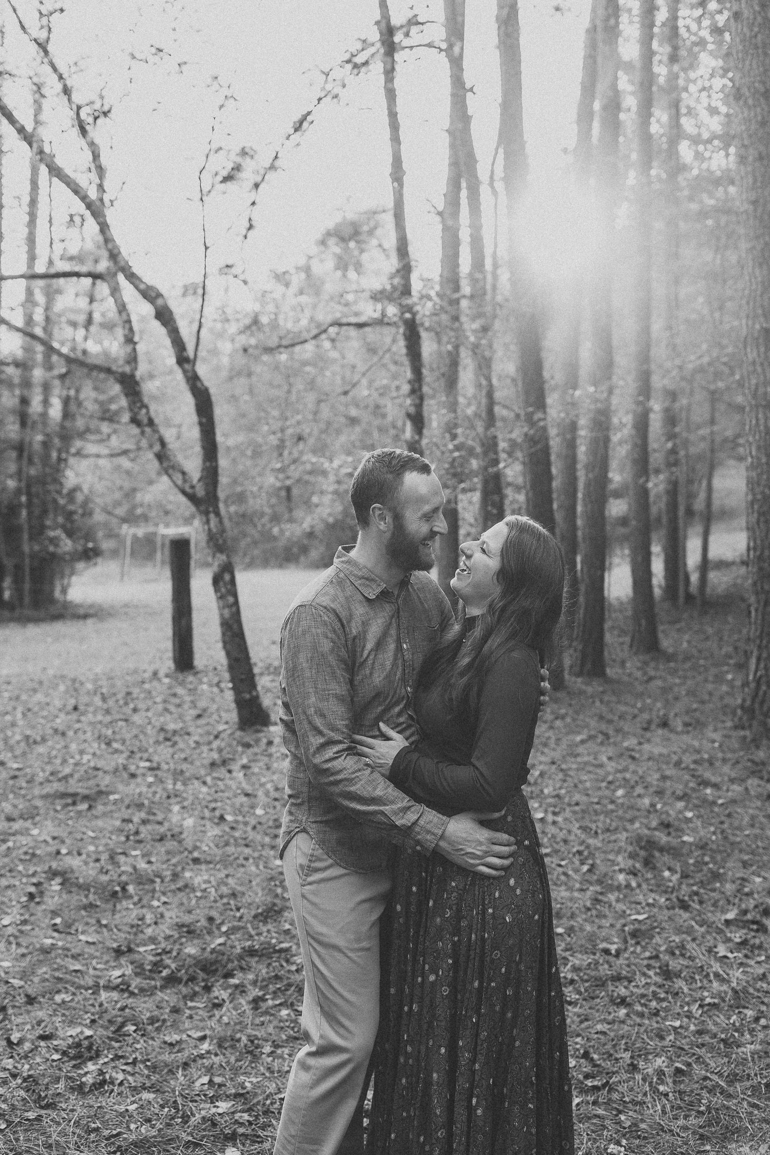 Black and white photo of a couple embracing and smiling in a sunlit forest during an outdoor engagement session.