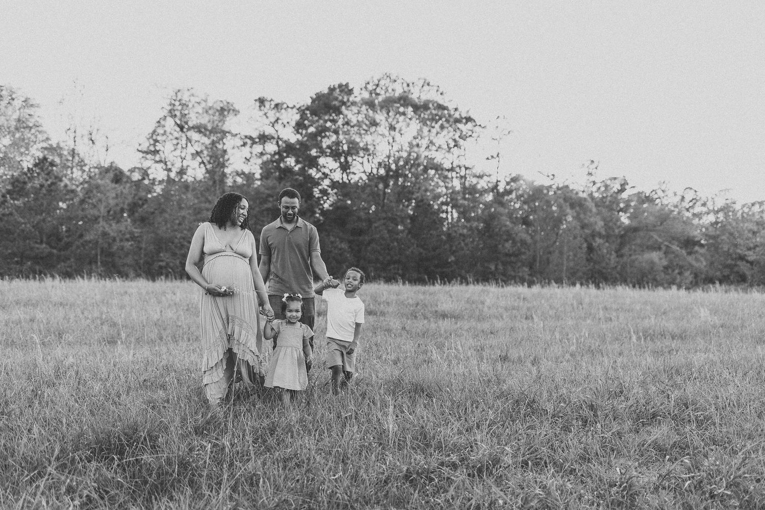 Black and white family portrait of pregnant mother, father, and two children walking in an open field.