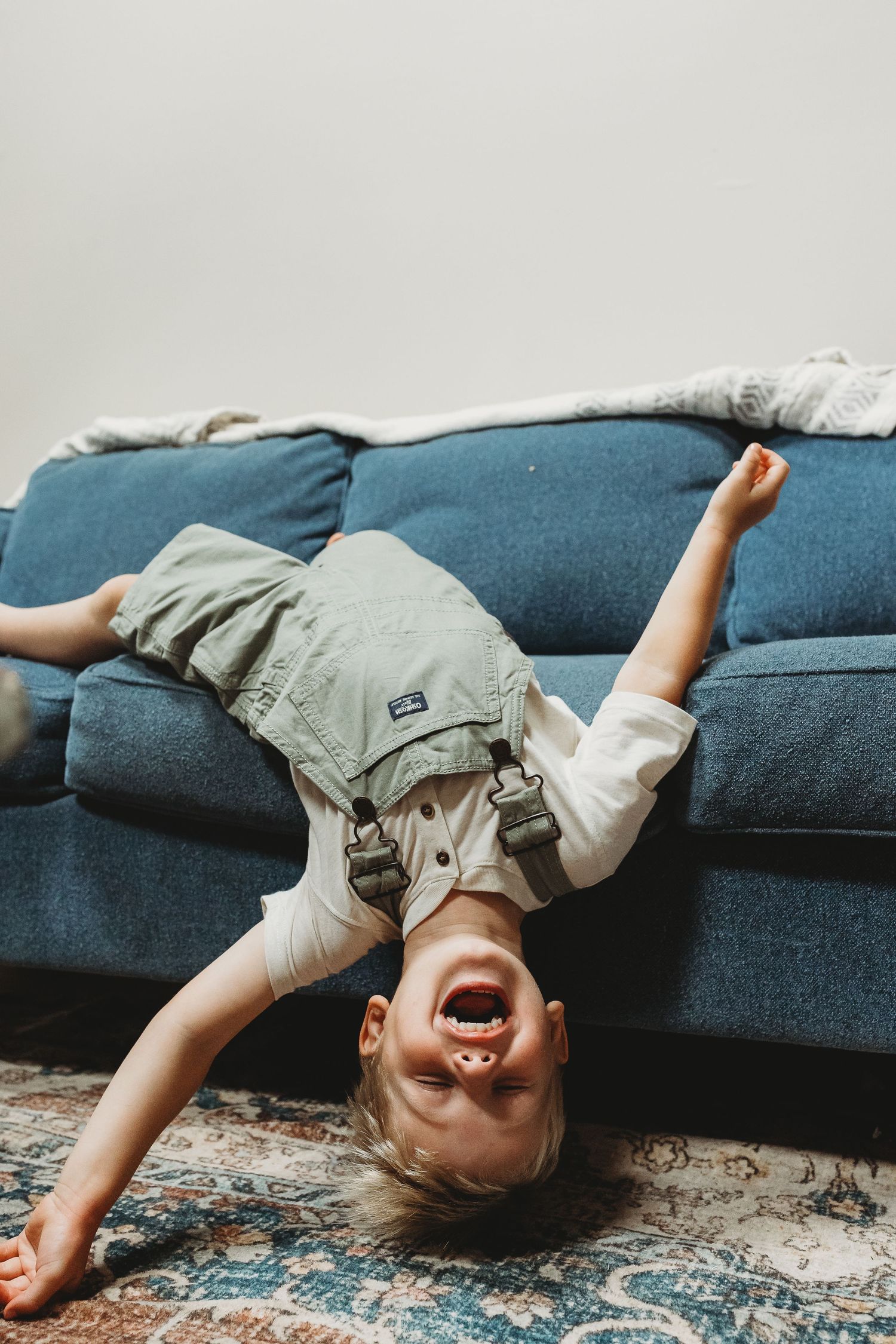 Young child in overalls laughing upside down on a blue couch, sliding onto a patterned rug.