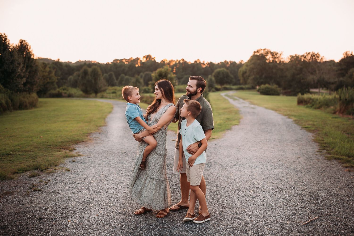 Family of four posing on a gravel path at golden hour, surrounded by lush green trees and fields.
