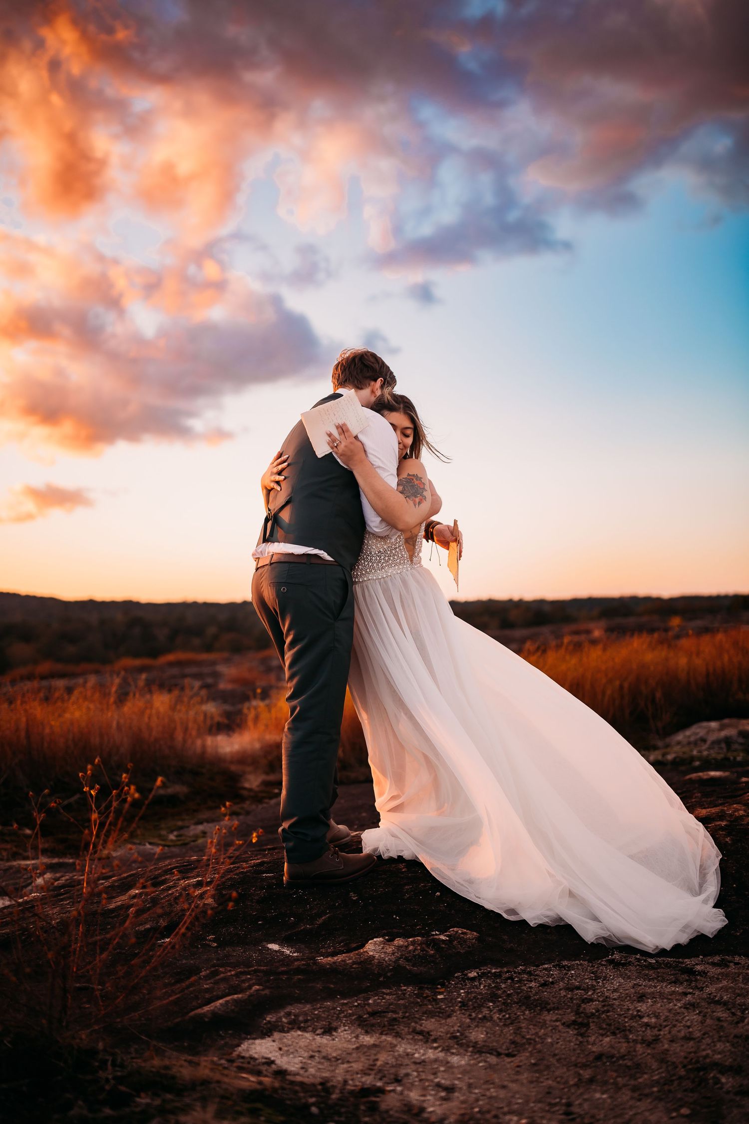 Bride and groom embrace at sunset with dramatic pink and blue clouds in an outdoor field wedding photo.