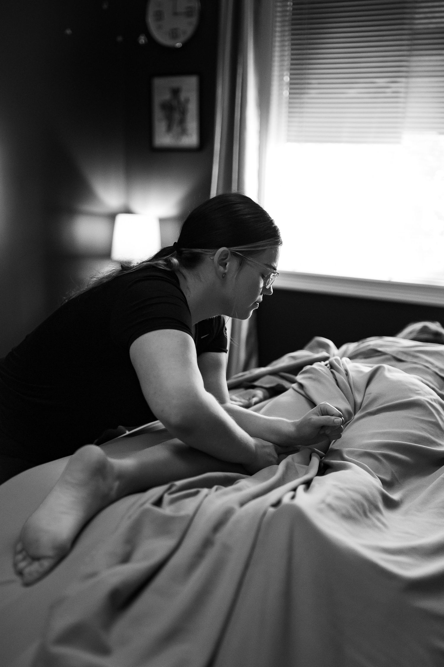 Black and white photo of woman leaning over bed in dimly lit bedroom.