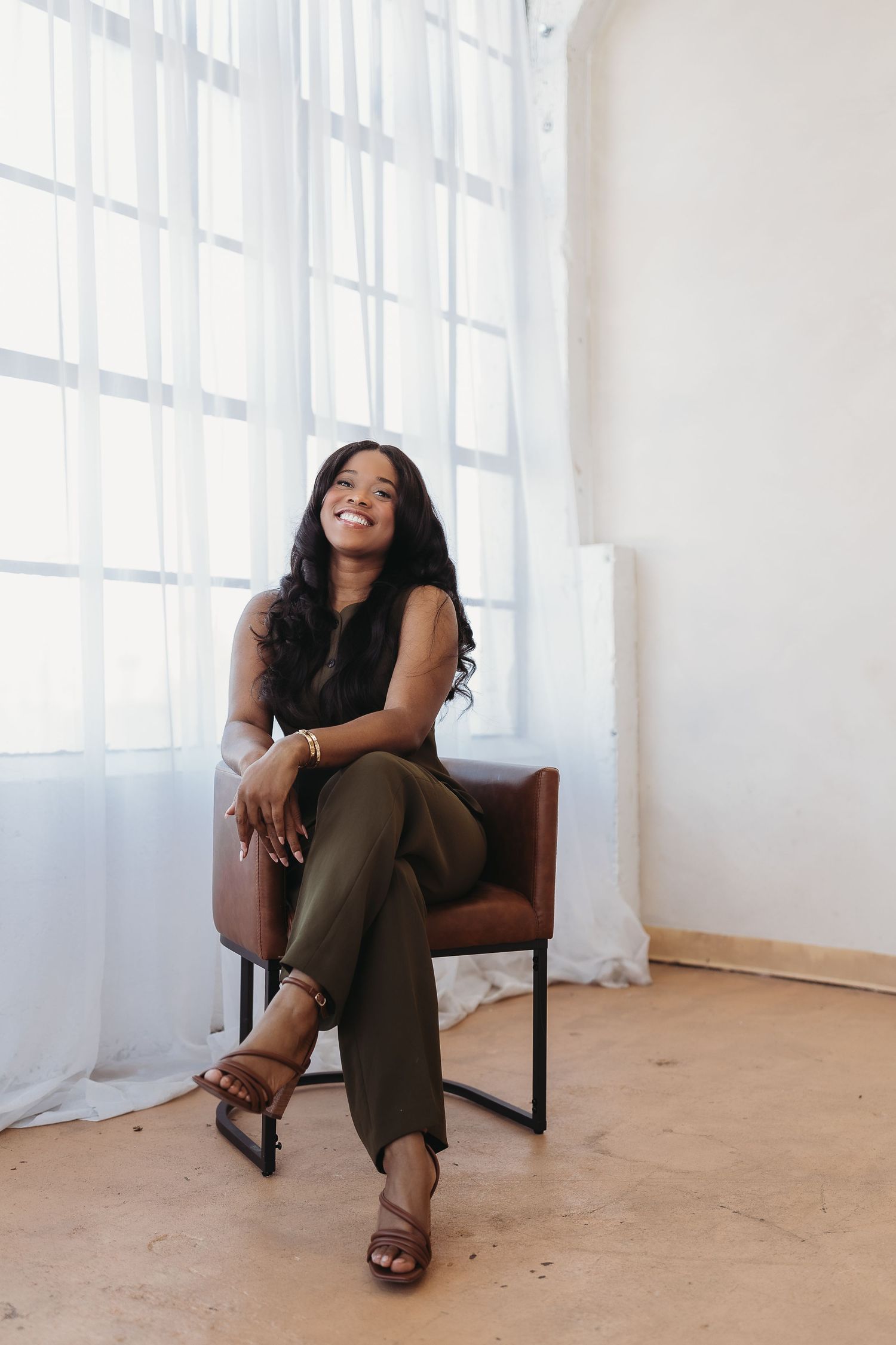 A smiling woman in a black sleeveless jumpsuit sits confidently in a brown leather chair by a bright window.